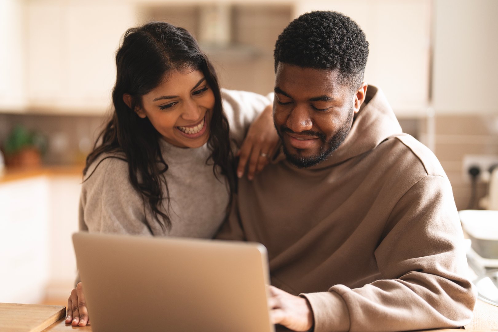 A couple shares a joyful moment in their bright kitchen, engaged with a laptop as they smile and interact in a warm, cozy atmosphere during a chill afternoon.