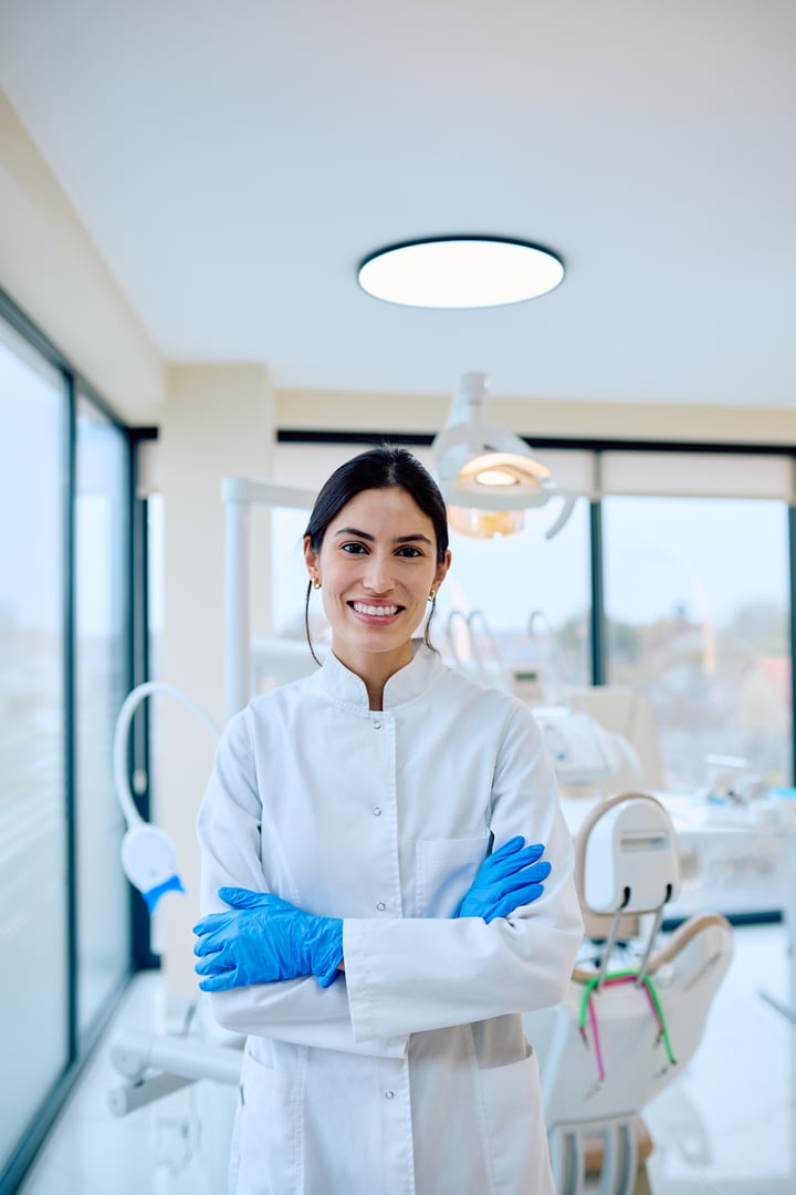 Female dentist wearing white uniform and blue gloves, smiling and posing with arms crossed in a modern dental clinic