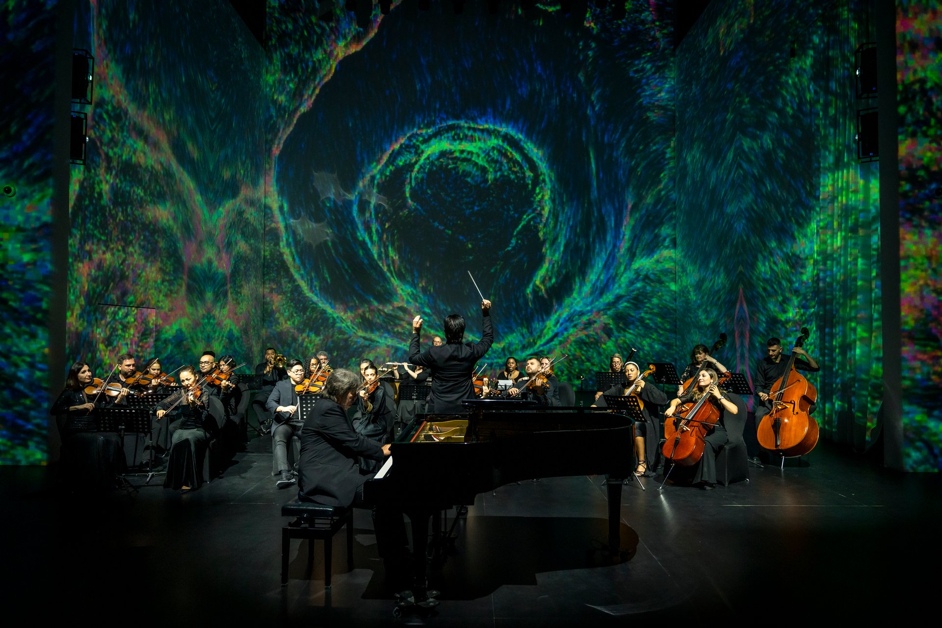 Orchestra performing on stage with a pianist and colorful swirling backdrop. Conductor at center leading musicians.