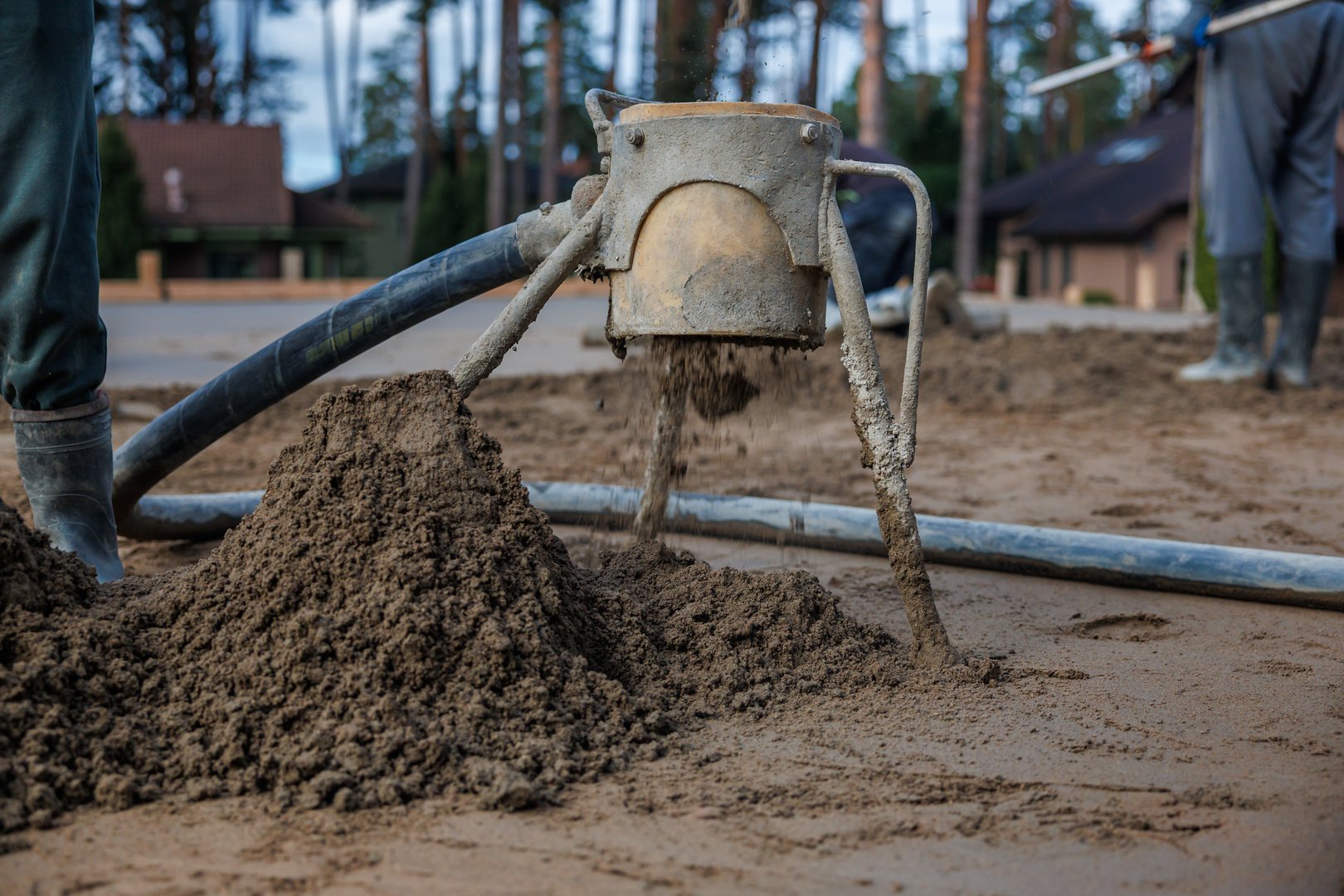 Concrete pouring tool dispenses wet concrete onto the ground as workers in boots operate nearby. Tall trees and sloped roof structures are visible.