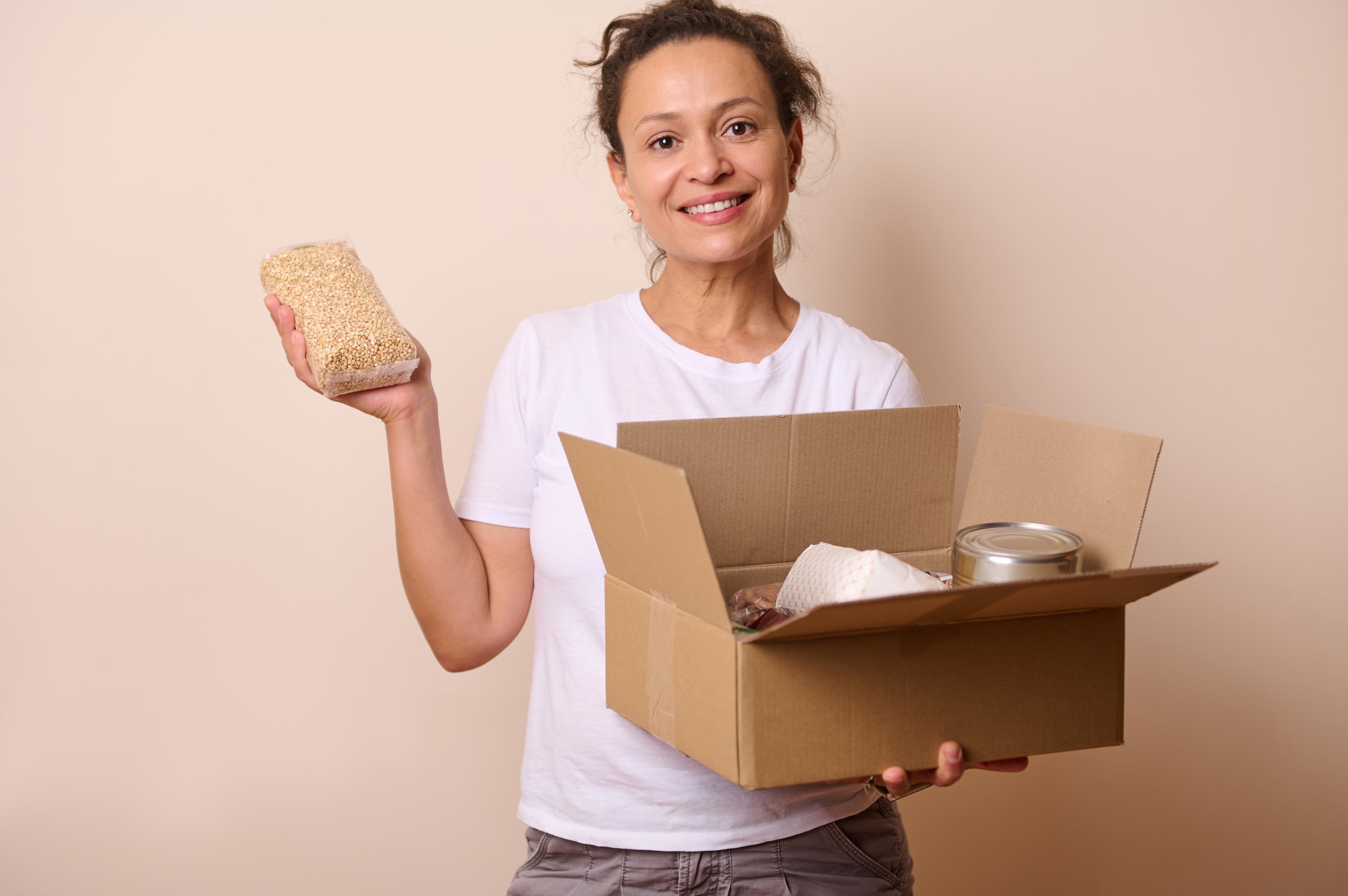 A cheerful woman holding a cardboard box with food items and a packet of grains. Represents kindness, charity, and support through donations.