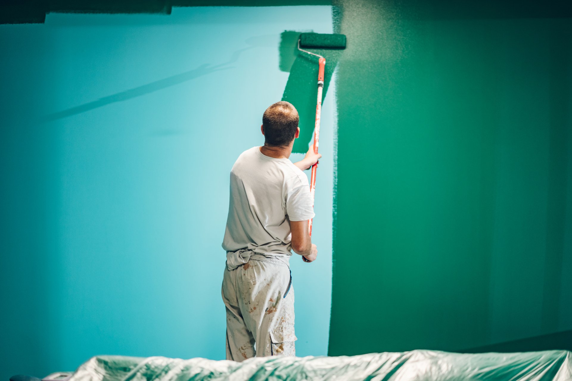 Painter applying green paint on a turquoise wall using a paint roller, seen from behind, during a home renovation project