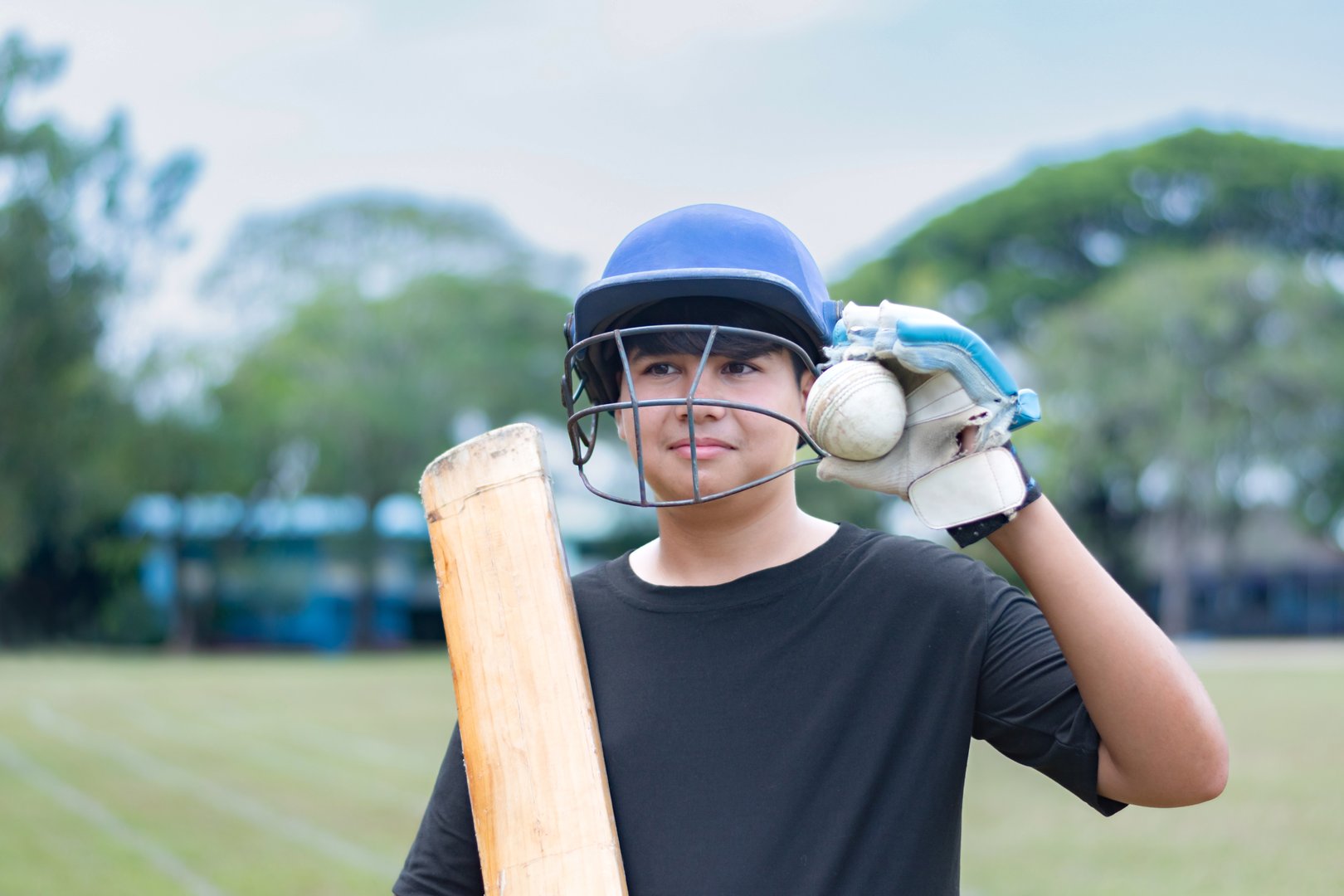 Young cricket sport player wears helmet and gloves, holding cricket bat and old cricket ball in outdoor field, soft focus, hobbies and education concept.