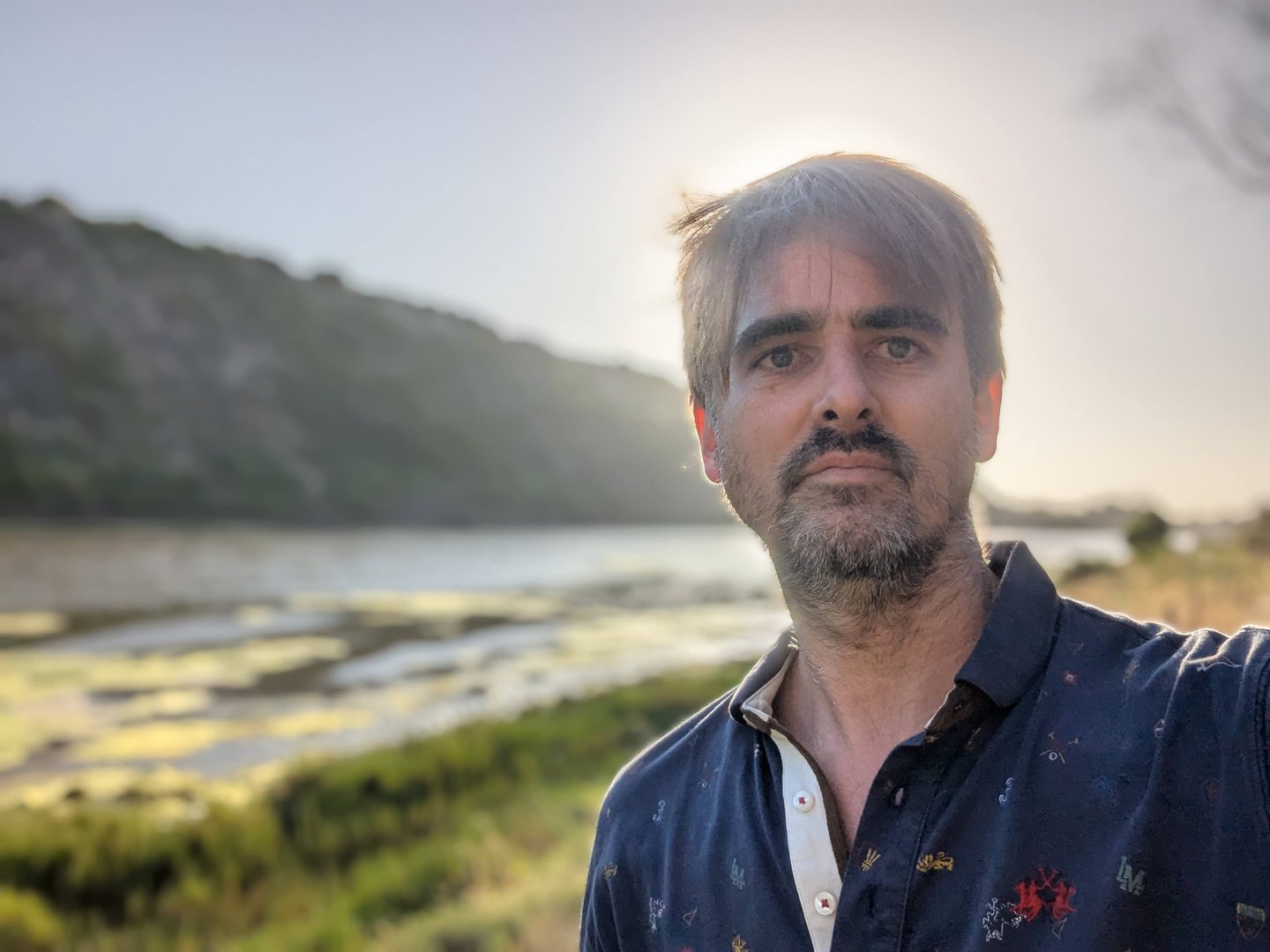 A man with gray hair and a beard stands outdoors near a body of water, with hilly terrain in the background at sunset.