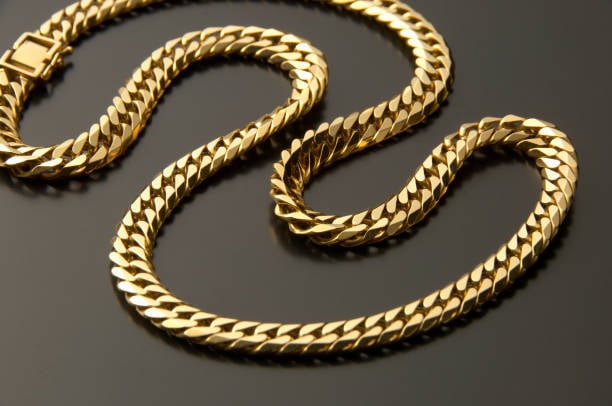 A gold chain necklace with a Curb link design placed on a glossy black surface, reflecting the necklace's polished finish.