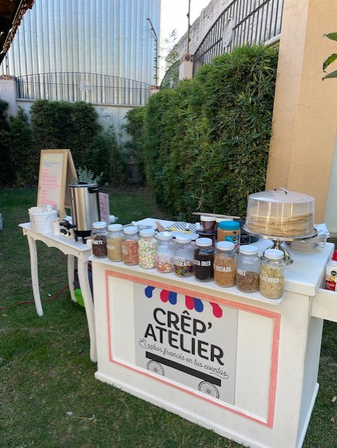 Outdoor crepe station with various toppings in jars, a hot beverage dispenser, and a chalkboard menu on a grassy area.
