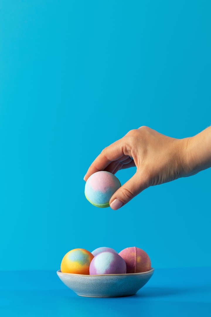 Woman takes colourful bath bomb from bowl put on bathroom counter on blue background. Bath bombs designed to fill bathtub with relaxing aroma and soothing bubbles