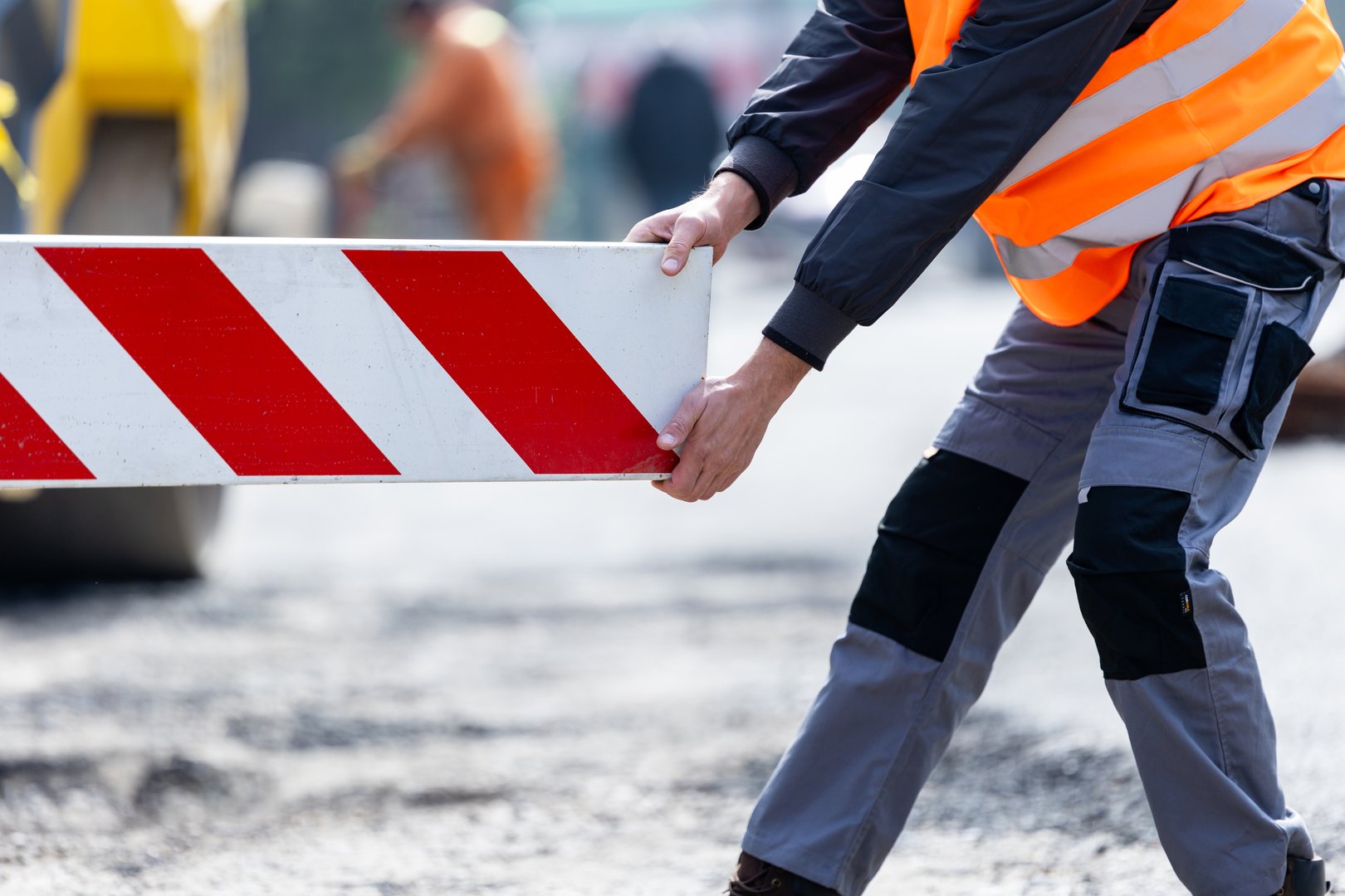 A construction worker in a high-visibility vest positions a safety barrier on a busy construction site, ensuring safe passage for vehicles and pedestrians.