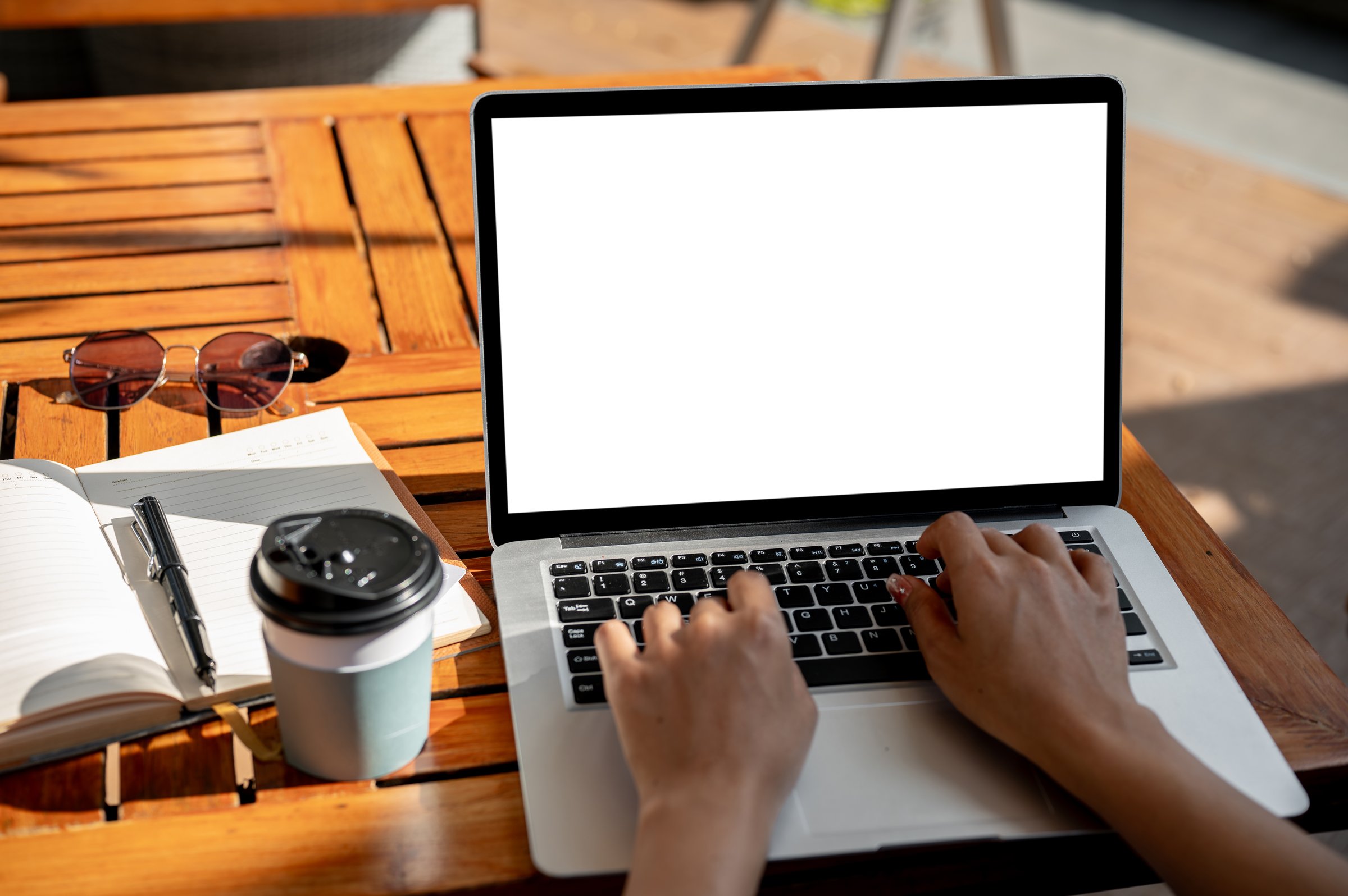 A close-up of a woman typing on her laptop keyboard while sitting at an outdoor table of a coffee shop on a sunny day in the city. the laptop with a white screen mockup