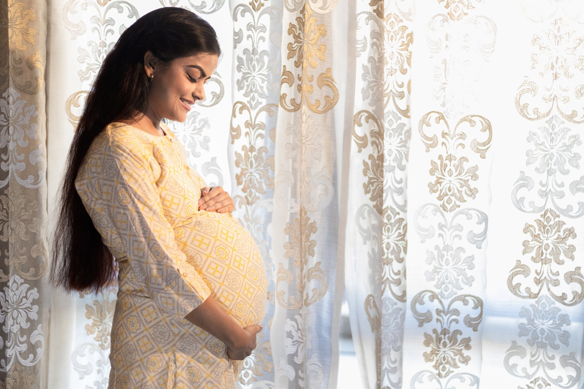 Happy pregnant indian woman touching big belly near window at home. people and expectation concept