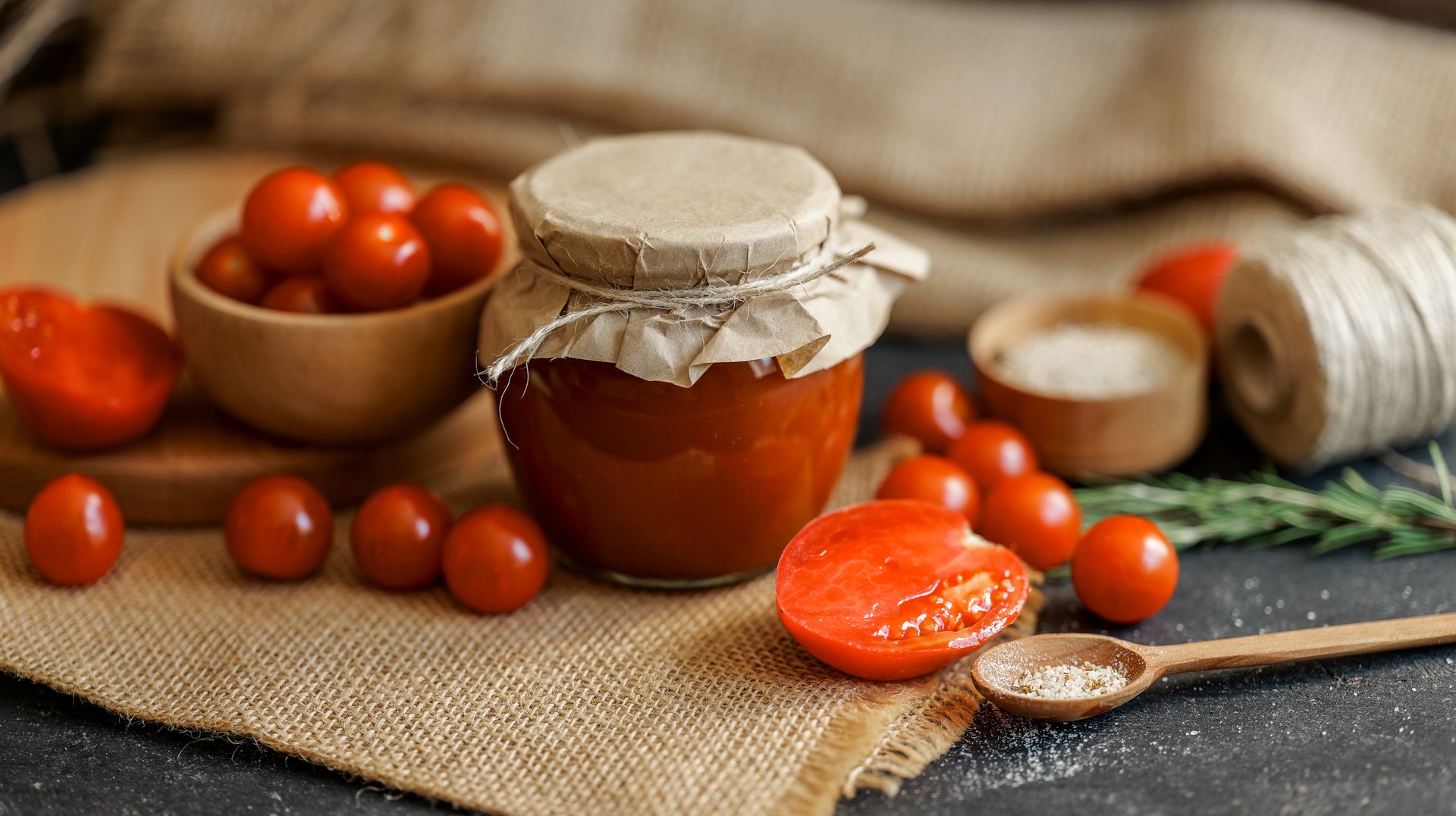 A jar of homemade tomato sauce is surrounded by fresh cherry tomatoes, herbs, and spices, creating a rustic and appetizing scene.