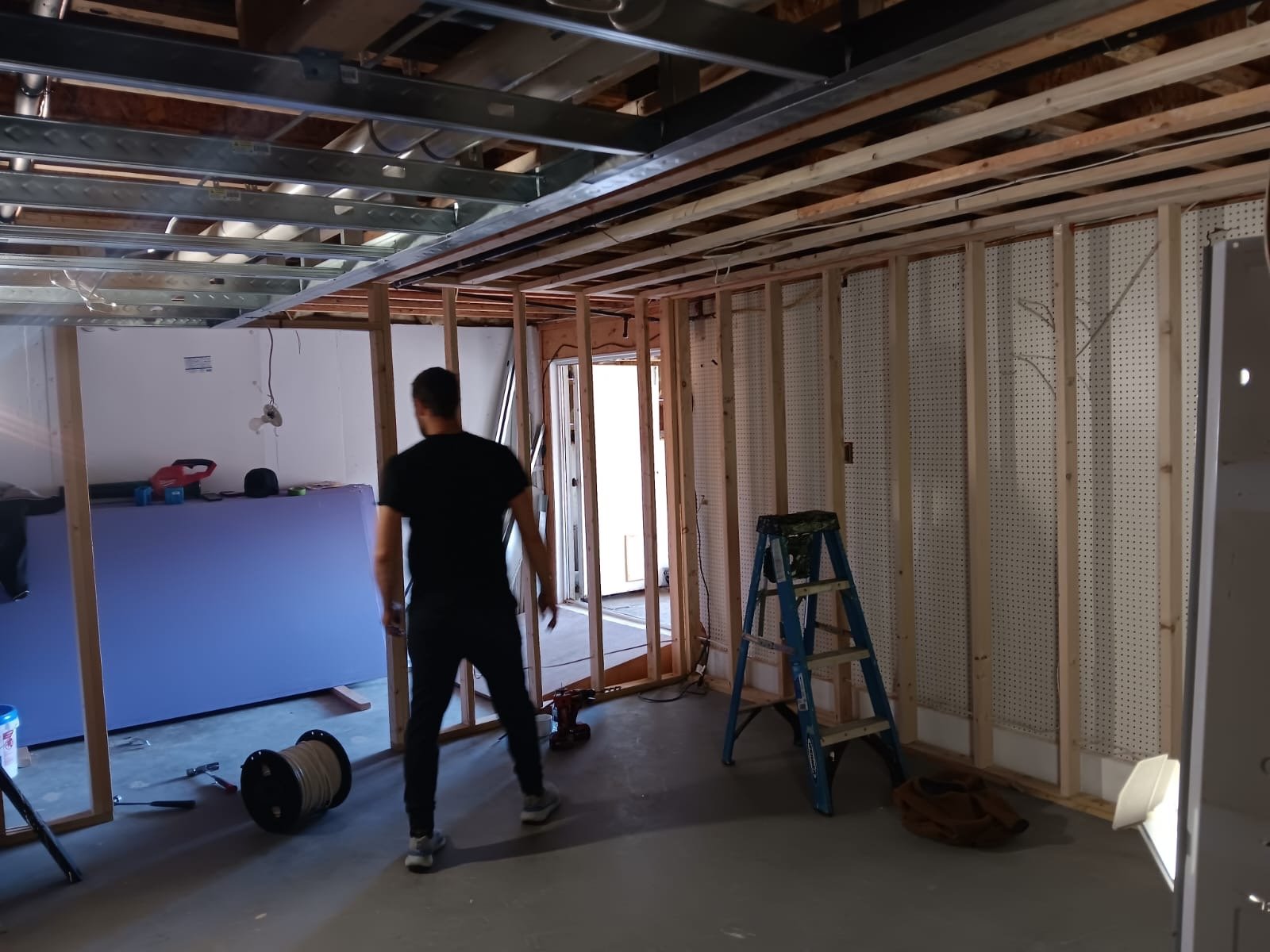 A man works on framing a room with exposed wood beams and drywall. A ladder and construction tools are visible.