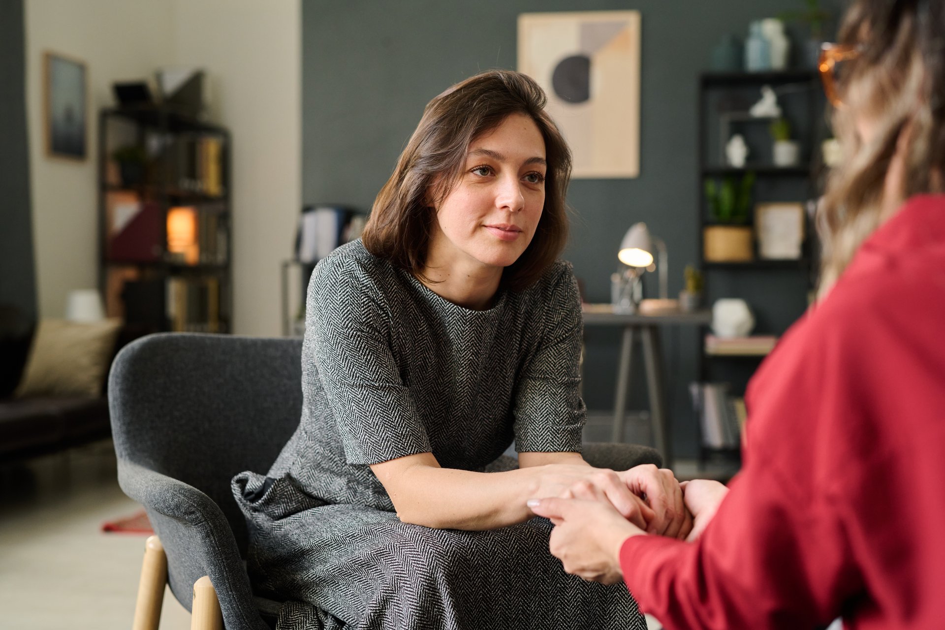 Female therapist sitting opposite to client, holding hands in supportive gesture during counseling session in well-decorated office environment