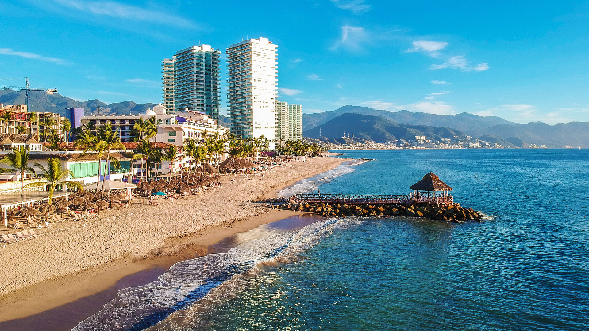 Sandy beach with high-rise buildings, tropical vegetation, and mountains in the background under a clear blue sky.