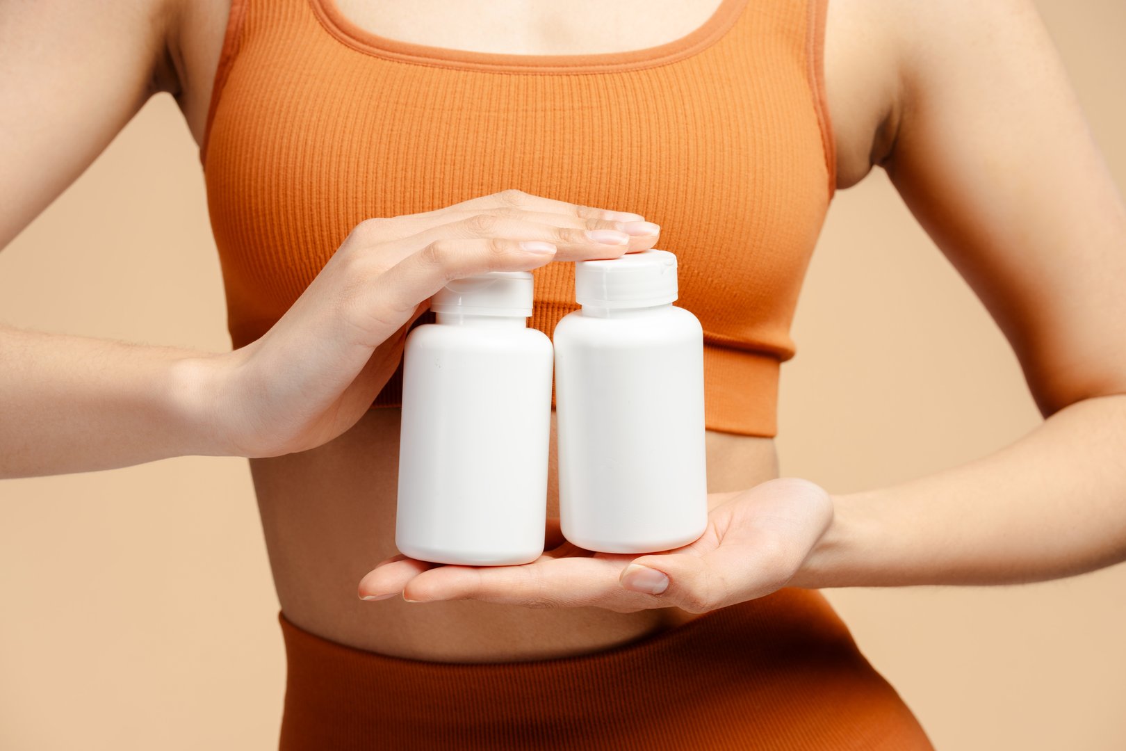 Closeup of athletic fitness woman in sportswear holding bottle with vitamins, capsules or pills and standing isolated over beige background. Healthy lifestyle and sport