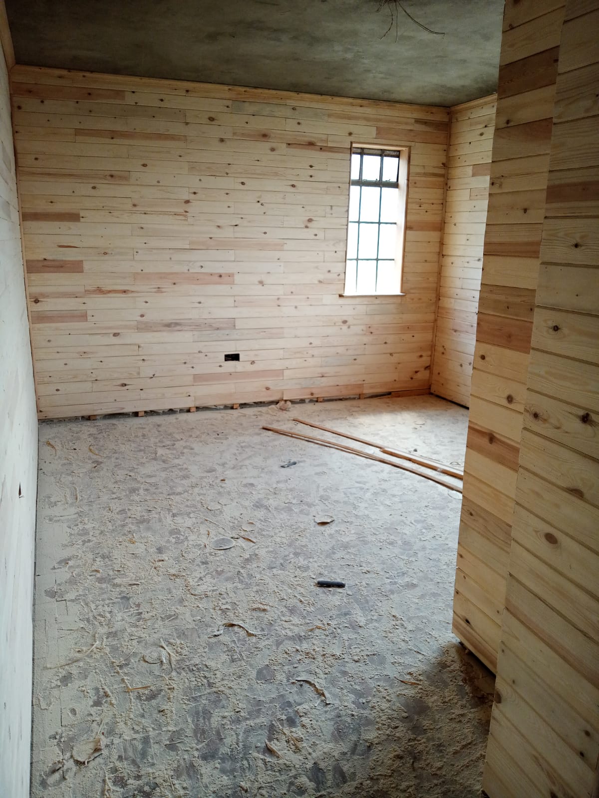An unfinished room with wooden paneled walls, concrete floor, and a single window letting in light.