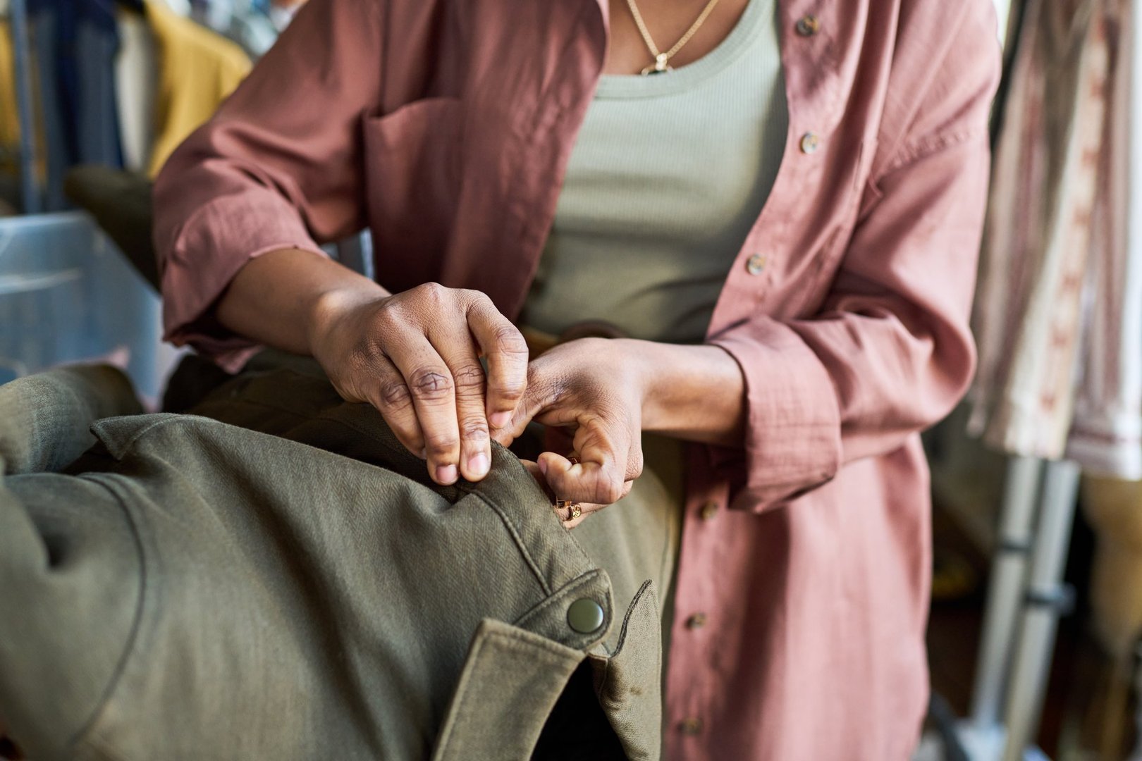 Young adult Black woman adjusting buttons on coat inside thrift shop, hands carefully fastening garment, clothing rack visible in background, focused on detailed work