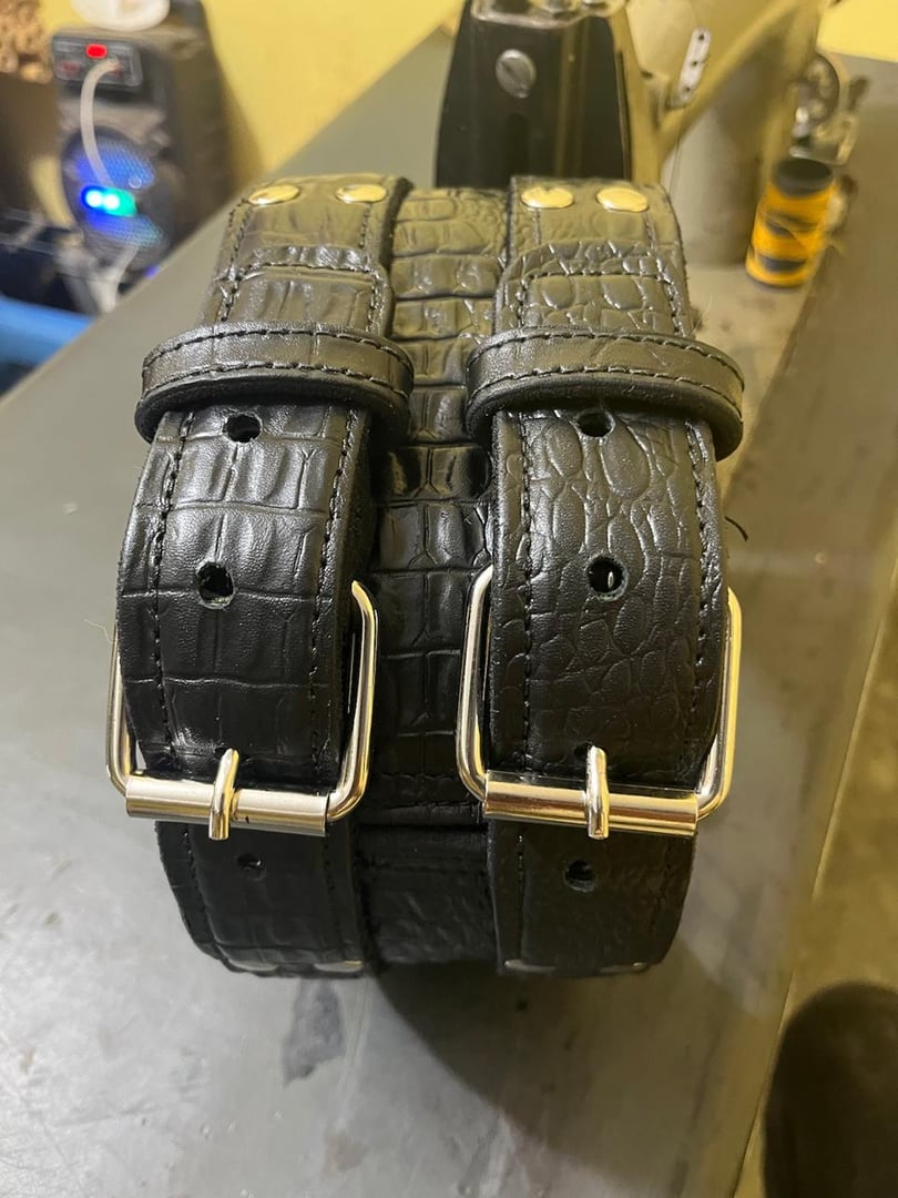 Two black leather belts with crocodile pattern and silver buckles placed on a table, with a sewing machine in the background.