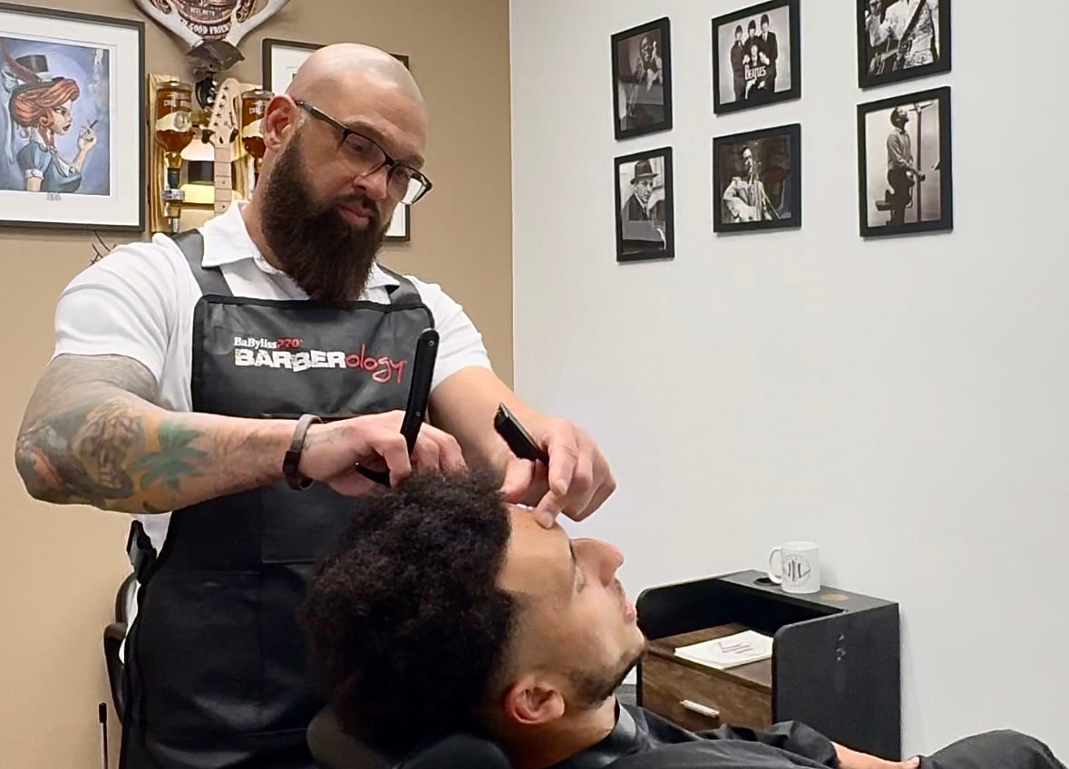 Barber shaping a man's hair with a comb and clipper in a shop, surrounded by framed photos on the wall.