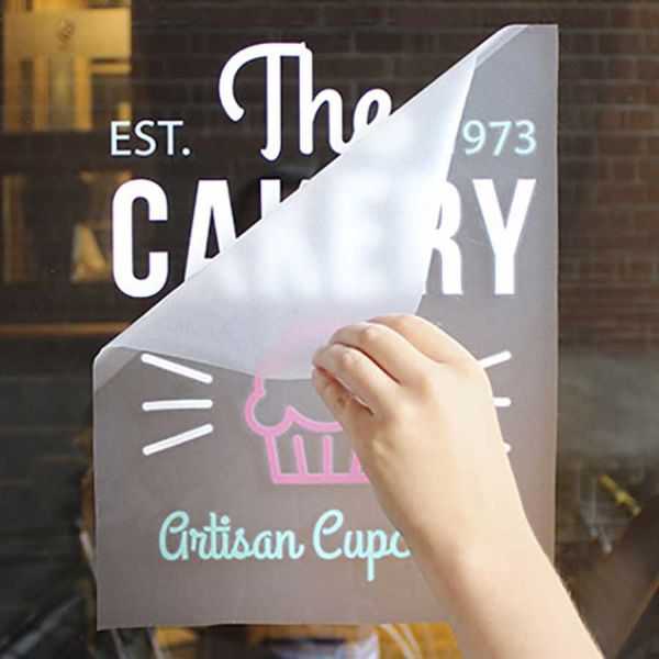 A hand applies a semi-transparent bakery sign to a window, displaying The Cakery and Artisan Cupcakes with a cupcake icon.