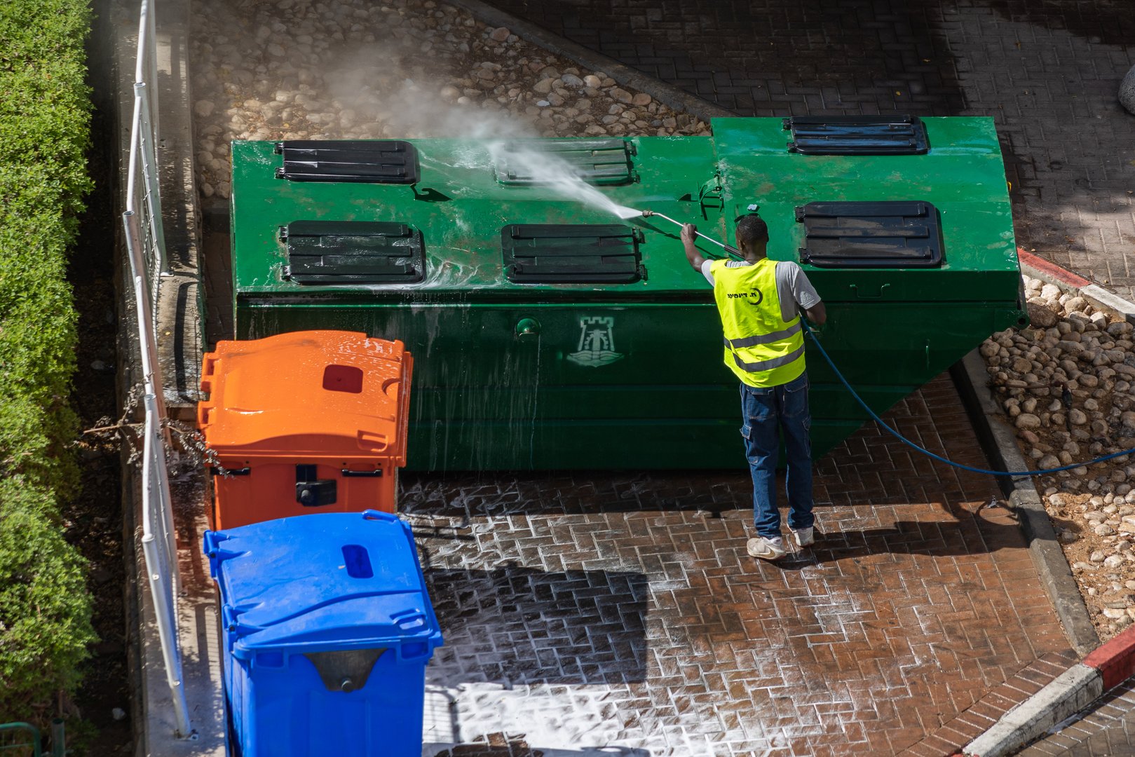 Eilat, Israel - July 9, 2024: Municipal worker in high visibility vest sinitizes garbage cans area. Different colors trash containers for sorting garbage. Cleaning company's name in hebrew on man's vest
