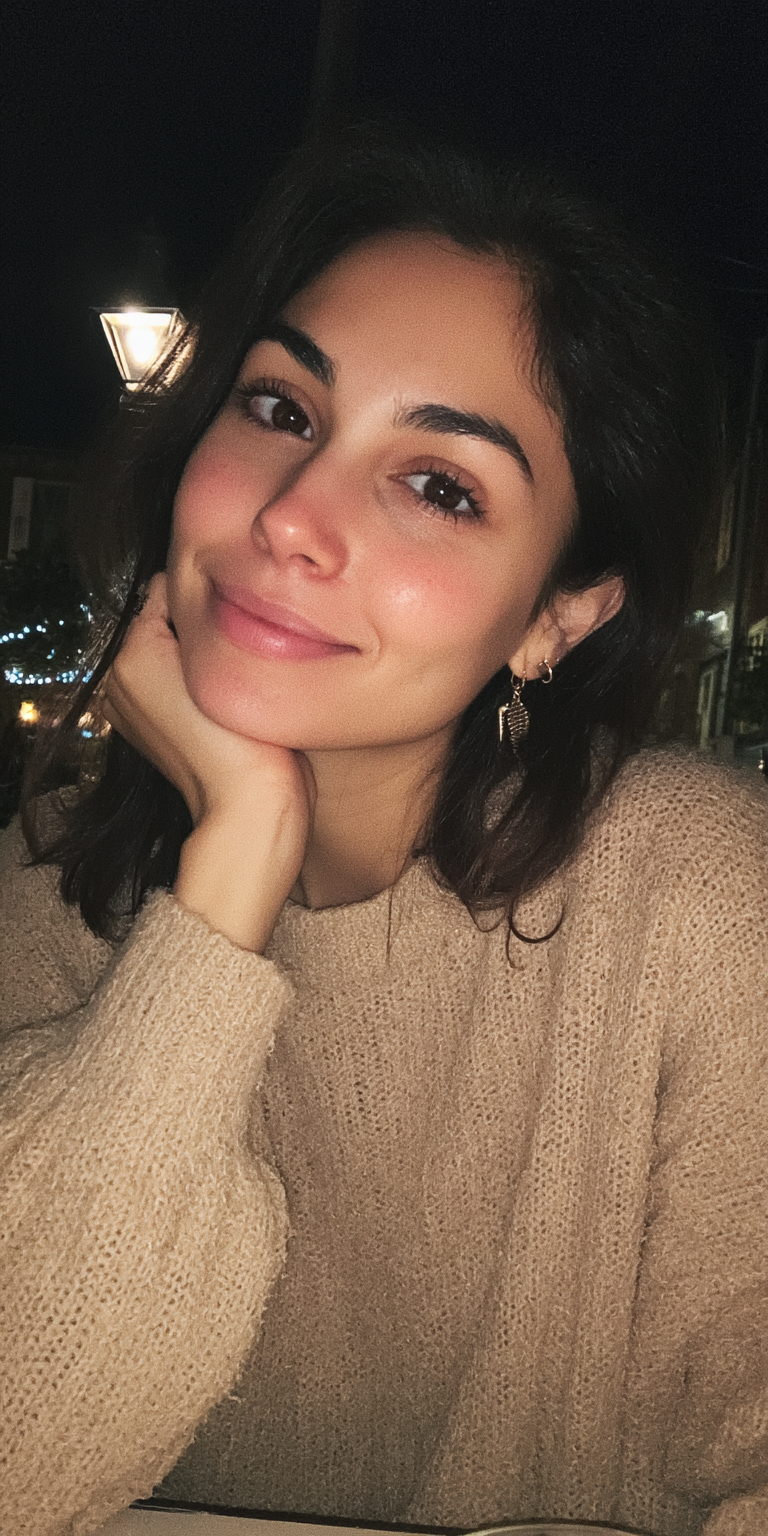 A woman with long dark hair, wearing a beige sweater, smiling gently with her hand on her chin, sitting outdoors at night.