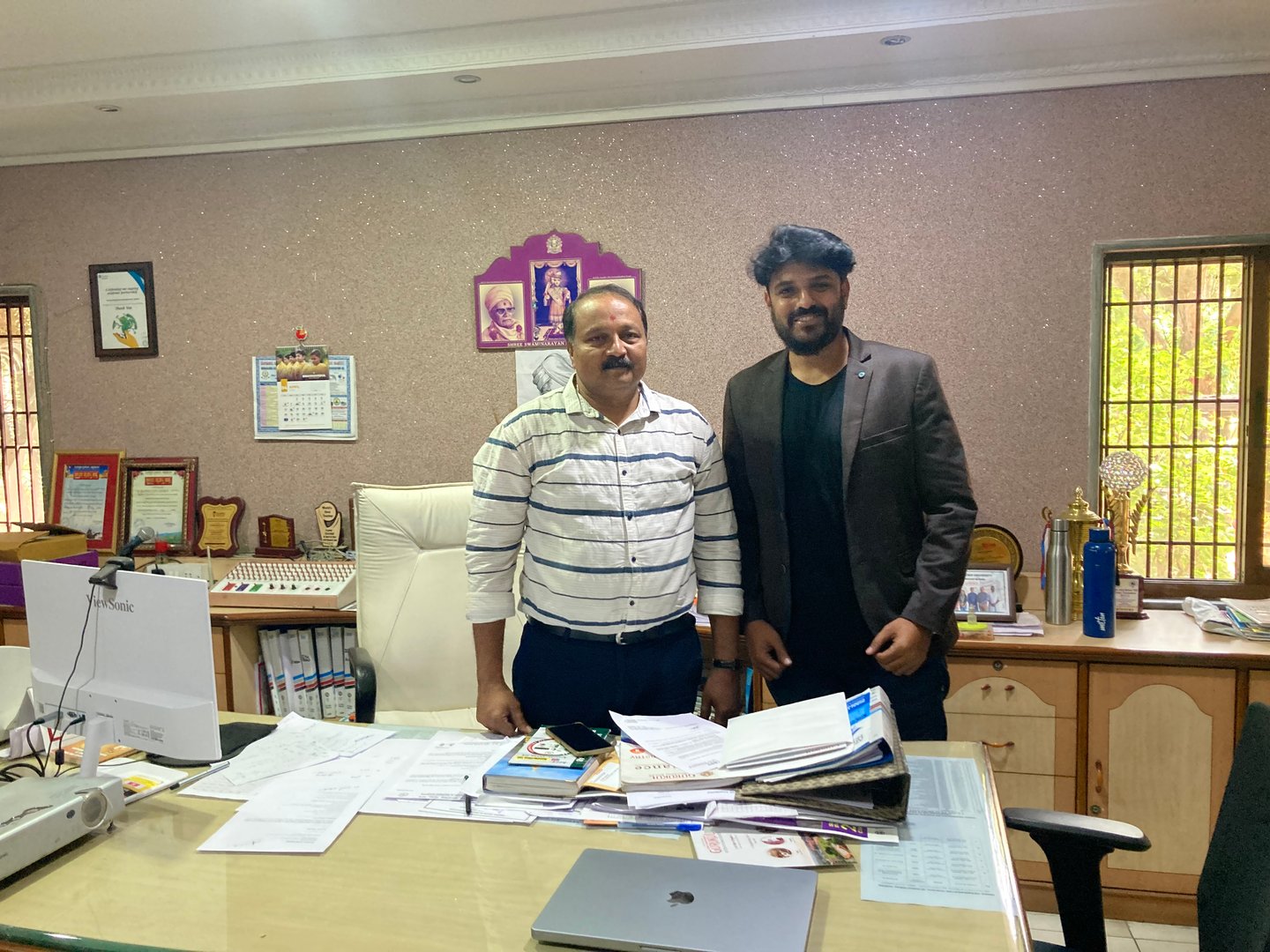 Two men standing in an office, surrounded by papers and awards on the desk. The room has framed pictures and certificates on the wall.