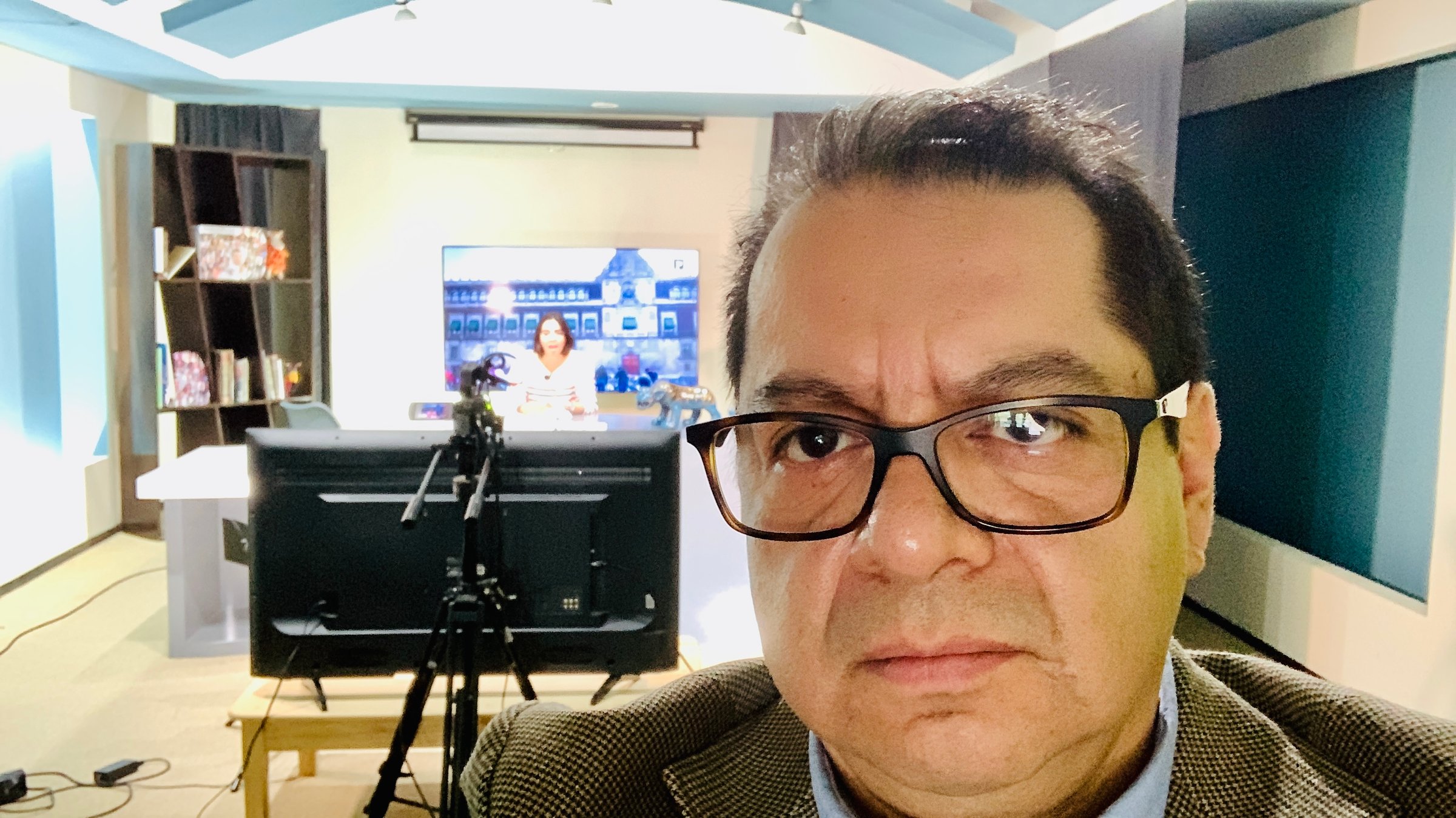 Man with glasses taking a selfie in a studio with a blurred screen and equipment in the background.