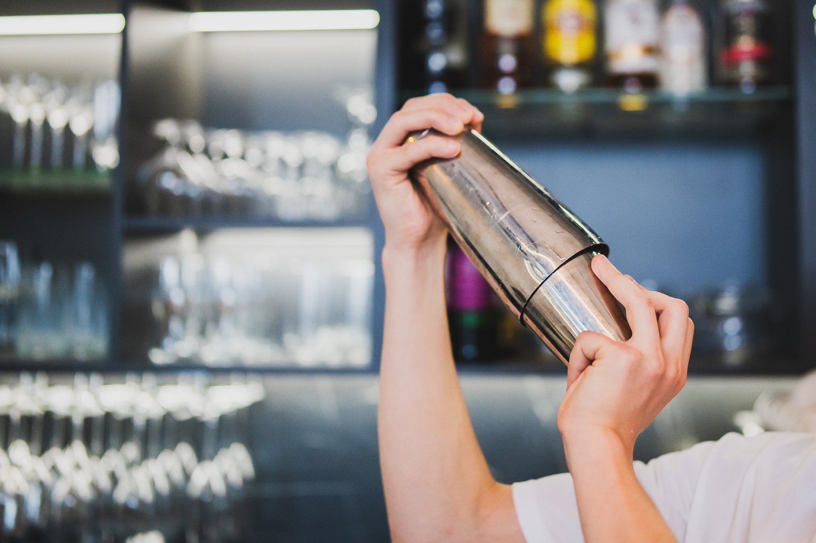 Professional bartender shaking a cocktail behind the bar counter with shelves of liquor bottles and clean glasses in the background, capturing dynamic bar atmosphere.