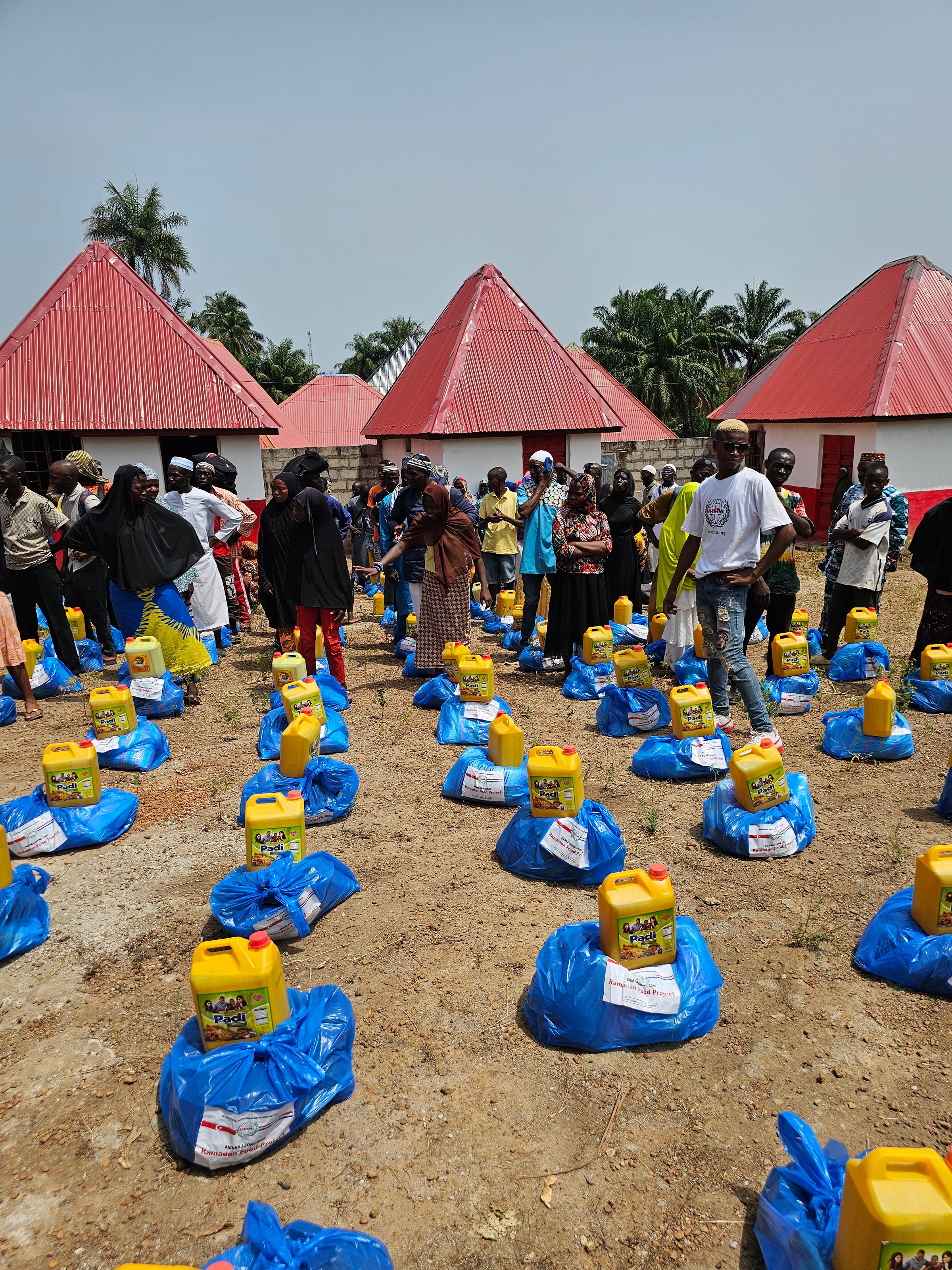 People distribute aid packages with blue bags and yellow containers in a rural setting with red-roofed buildings.