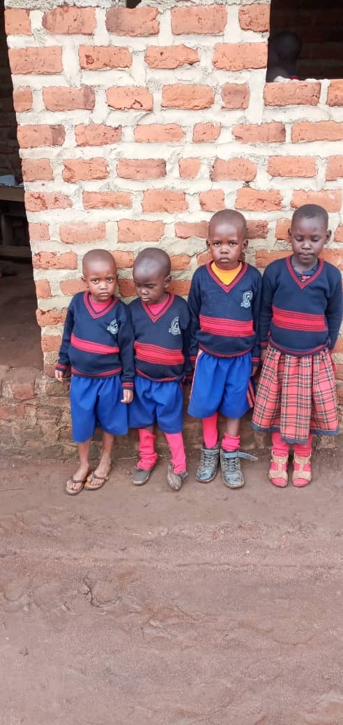 Four young children in blue and red school uniforms stand in front of a brick wall, smiling for the camera.