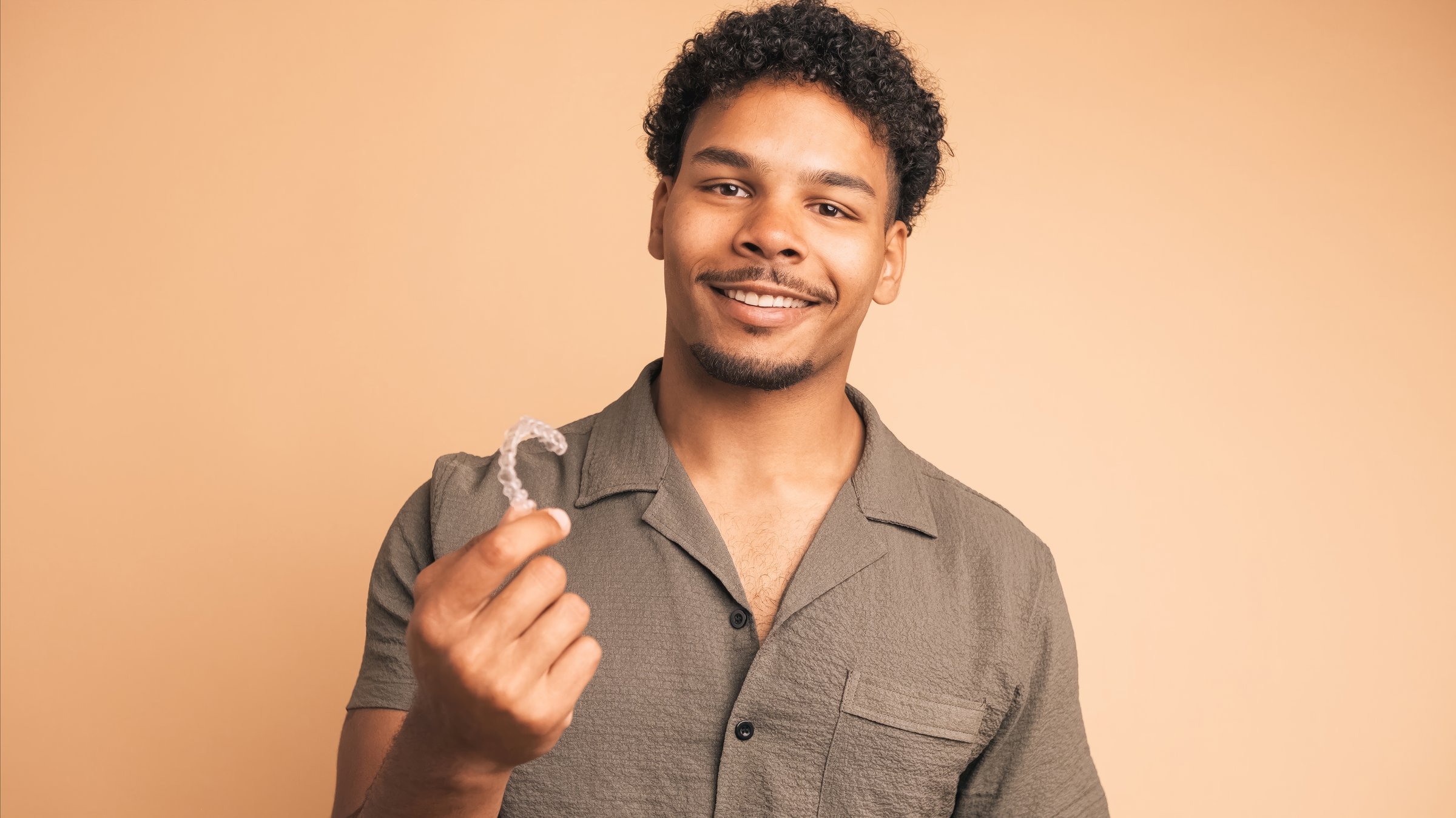 Happy young Brazilian male in shirt with afro hair showing invisible and removable clear aligner braces while looking at camera against beige background