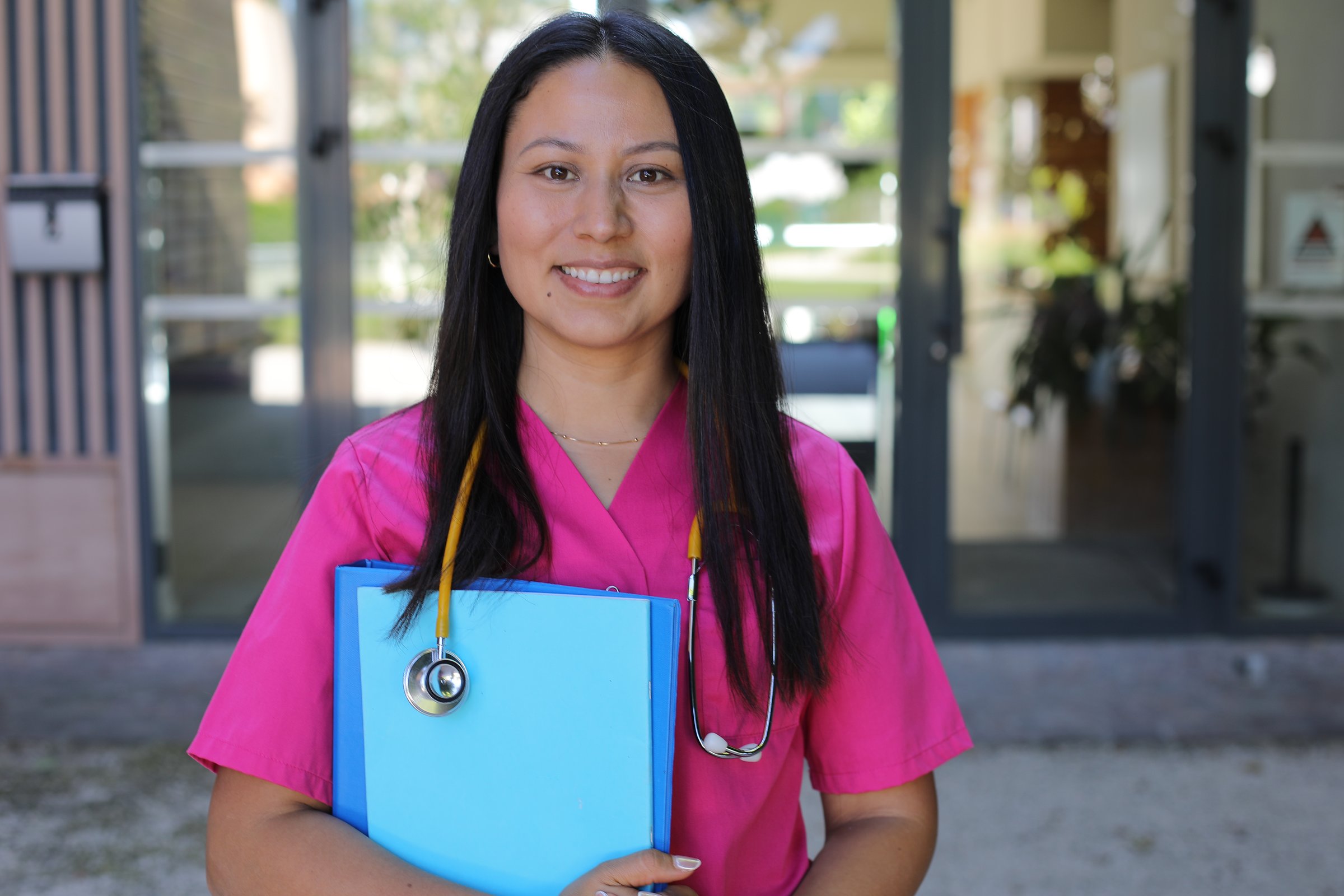 A young and beautiful Hispanic female healthcare worker is posing in a hospital entrance. She wears pink scrups and  yellow stethoscope.