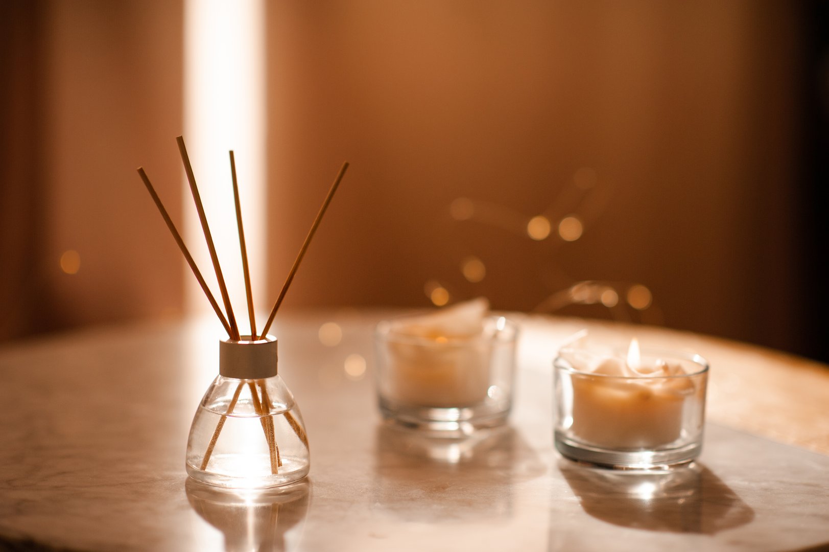 Home liquid perfume in glass bottle with bamboo sticks and scented candle on marble table over glowing lights in living room close up. Aromatherapy.