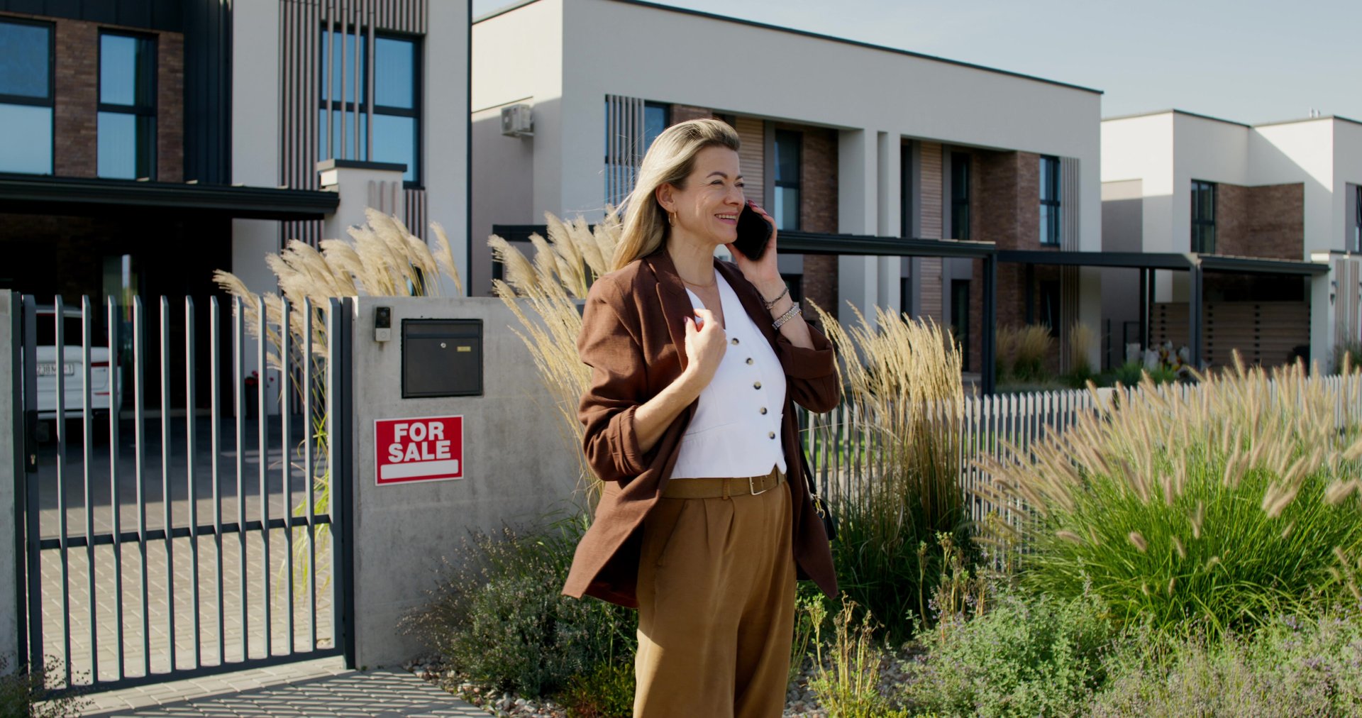 A stylish woman is talking on a mobile phone standing near a house for sale. She smiles. A sign for sale on the fence