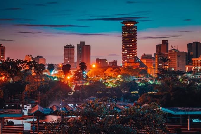 Cityscape at twilight with illuminated buildings, including a cylindrical tower, against a colorful sky.