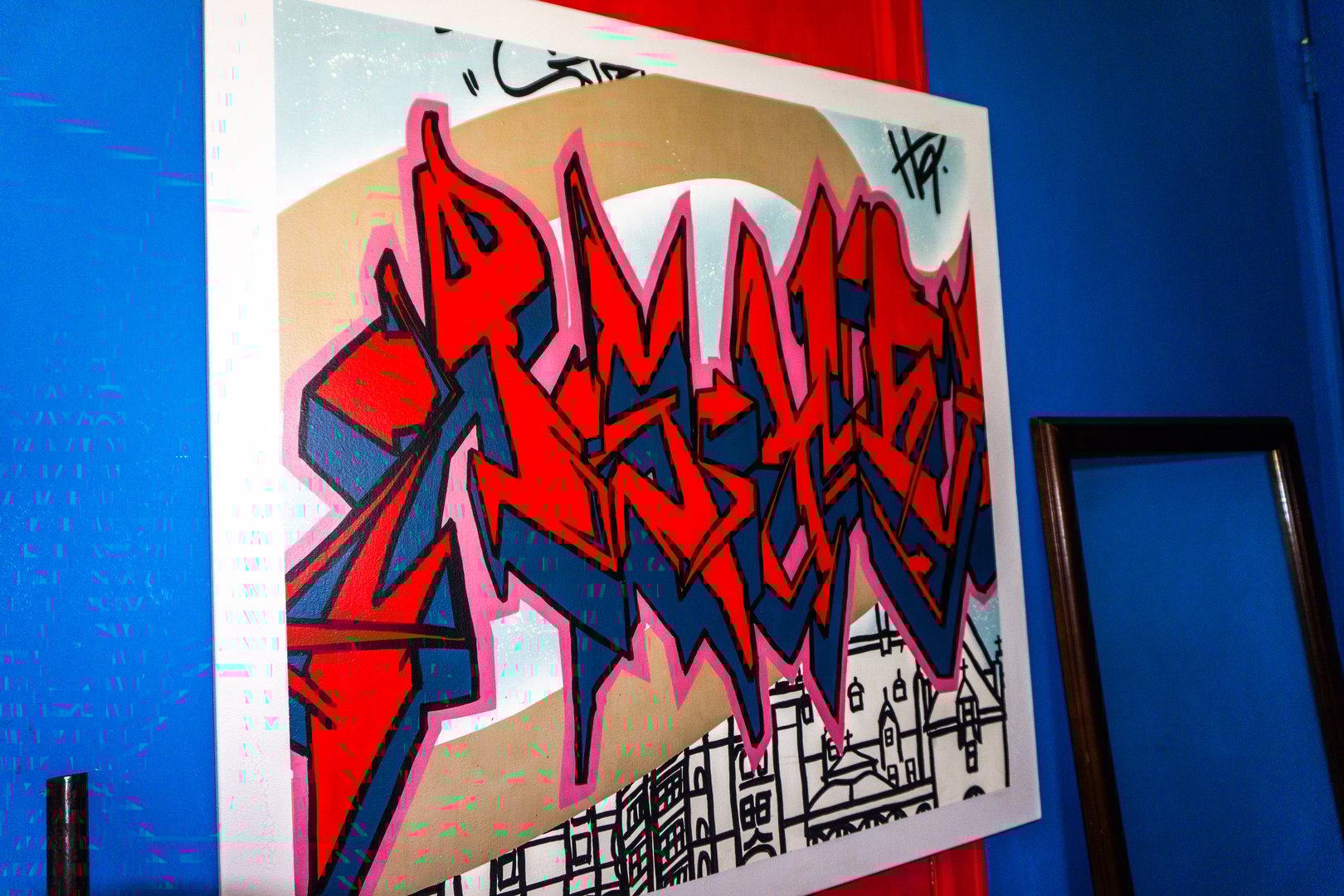 Colorful graffiti art with bold red and blue lettering on a framed canvas against a blue wall.