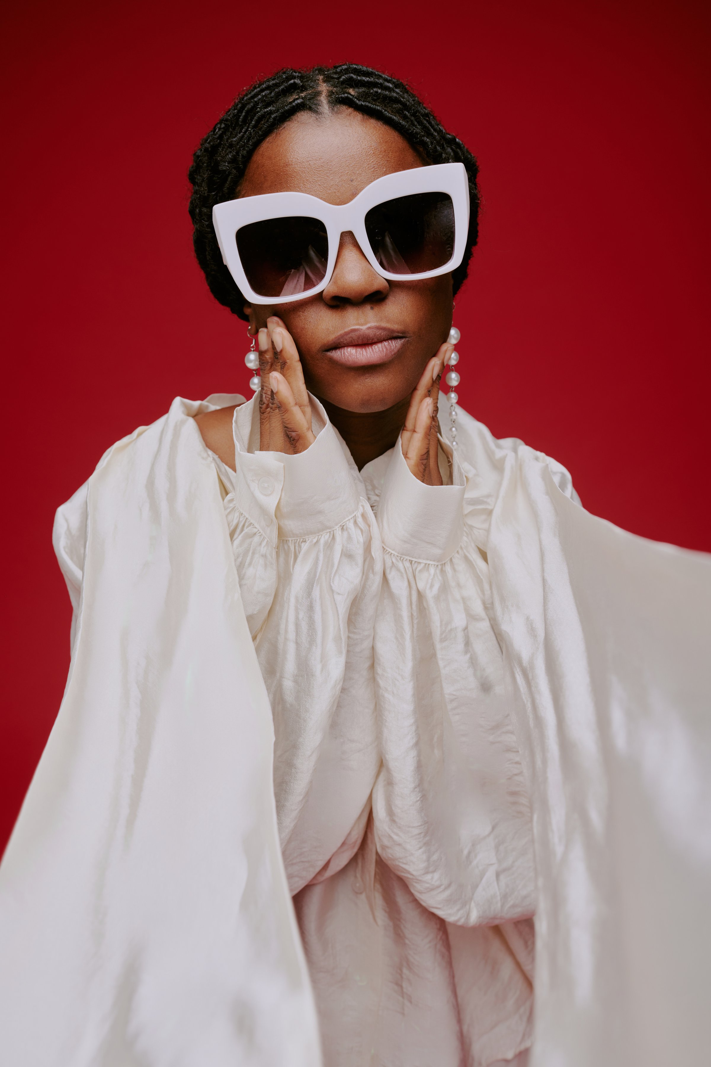 African American woman posing against red background, wearing oversized sunglasses and elegant attire. Displaying a confident expression and accessorizing with statement jewelry