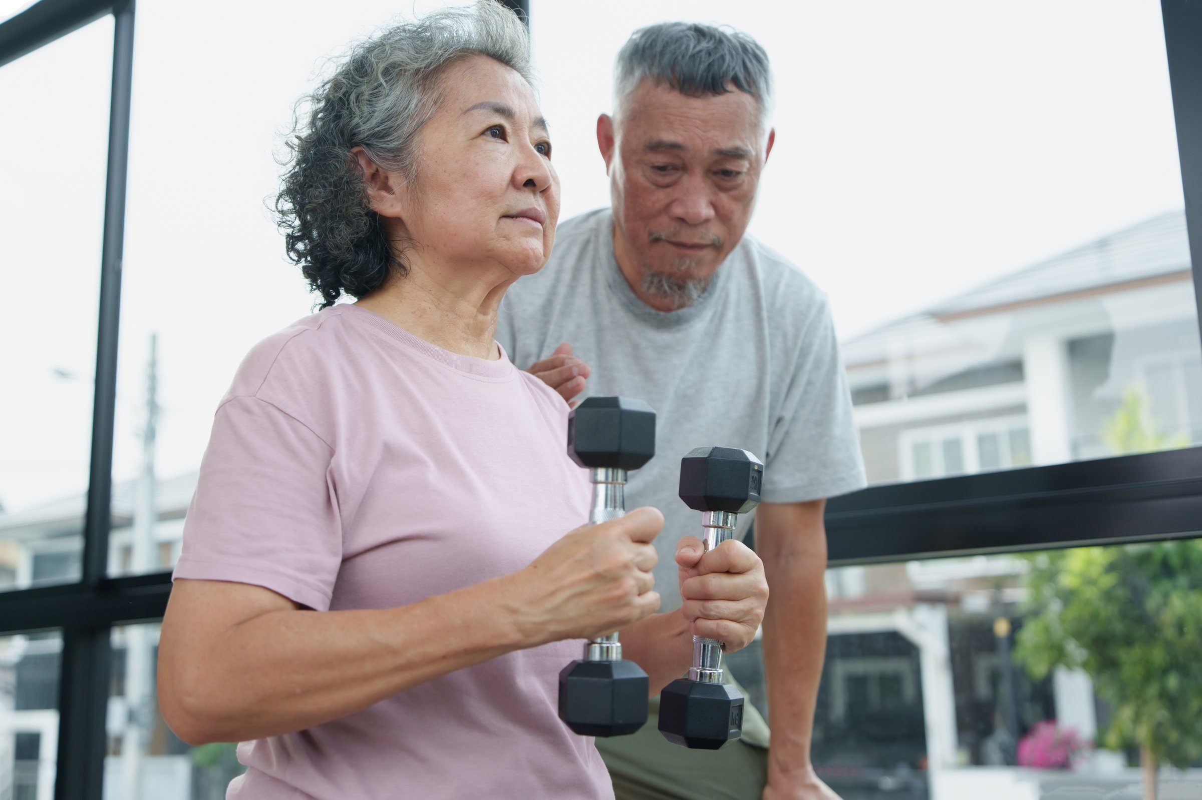 Asian Senior couple is working out together in a gym, focusing on stretching and strength exercises. Elderly Woman holds dumbbells while her husband encourages her, showcasing a healthy lifestyle.