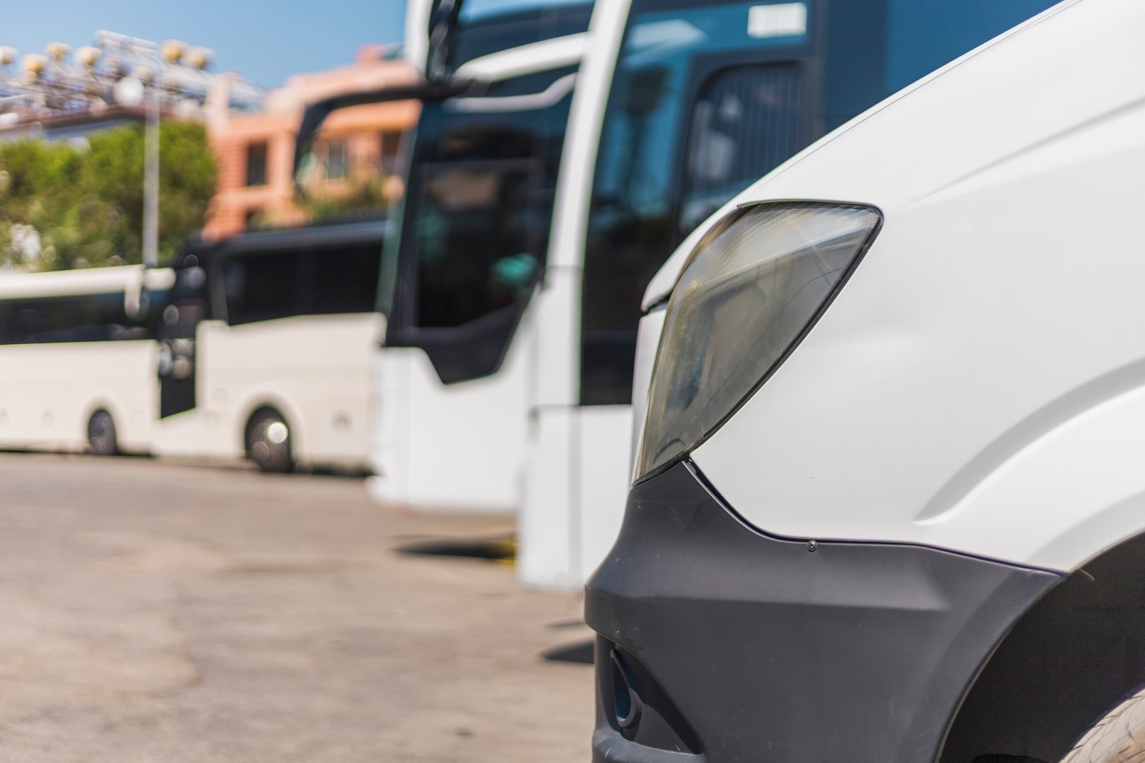 A close view of a white van shows its front while larger buses stand parked in the background. The scene is set at a busy transport station on a sunny day.