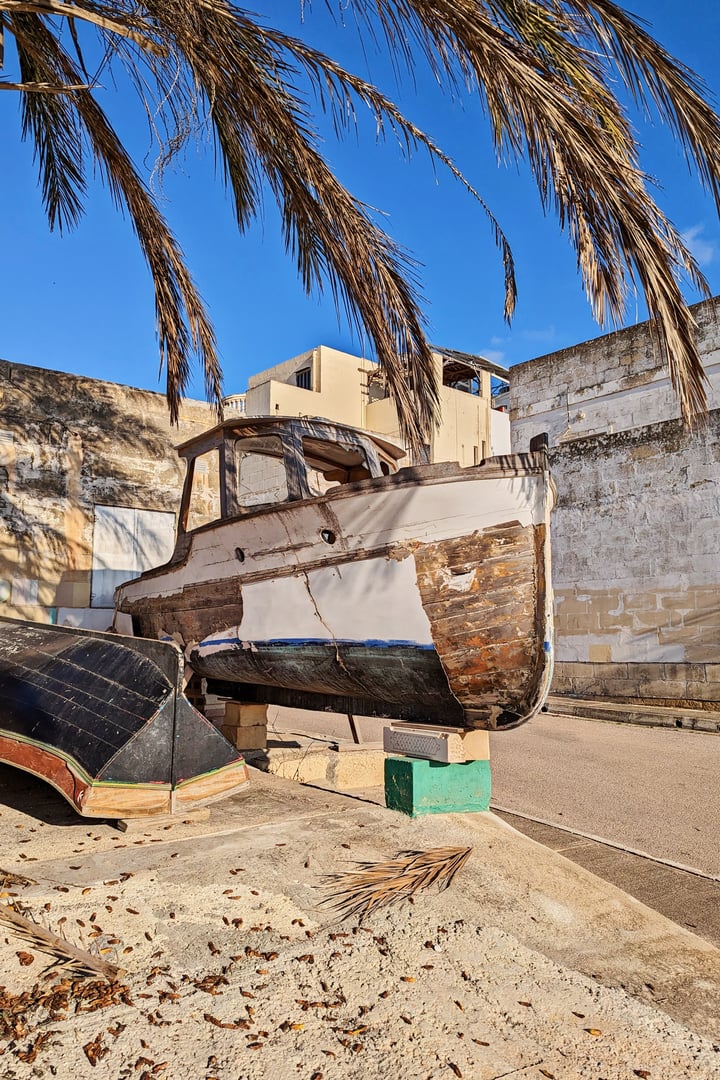 Rustic wooden boat with peeling hull sits under swaying palm leaves in coastal town, surrounded by historic stone walls