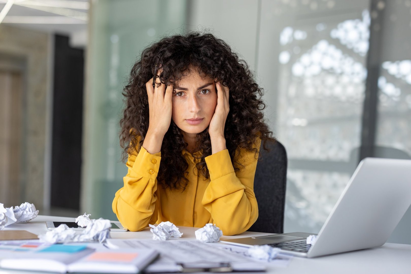 Stressed Latin American businesswoman in yellow shirt feels pressure at work, surrounded by crumpled papers and laptop. Concept of office stress, frustration, overwork.