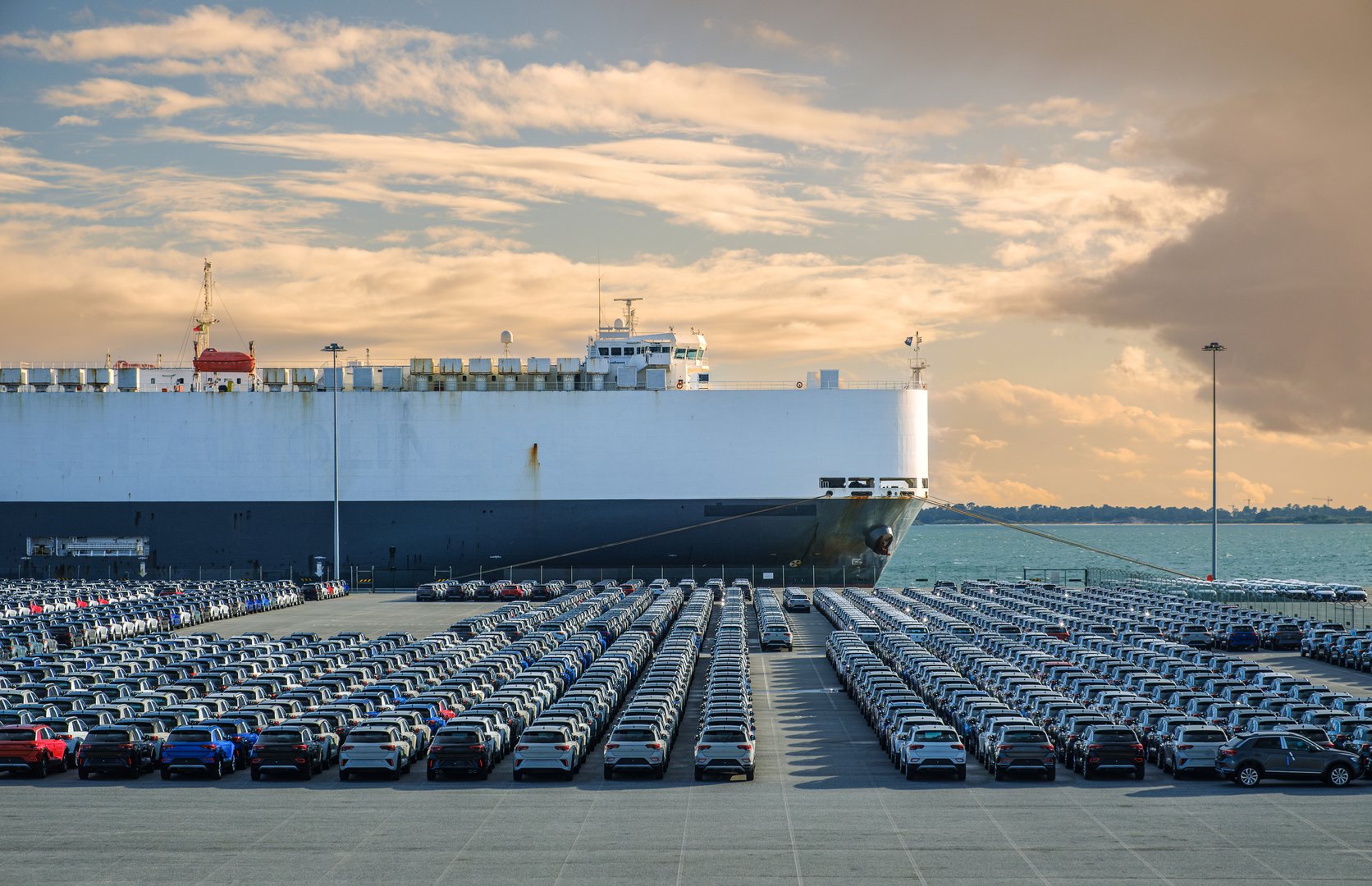 Large vehicle carrier ship known as RO-RO at port on sunny day, large parking area filled with cars. Blue sky and colorful clouds, vehicle transportation.