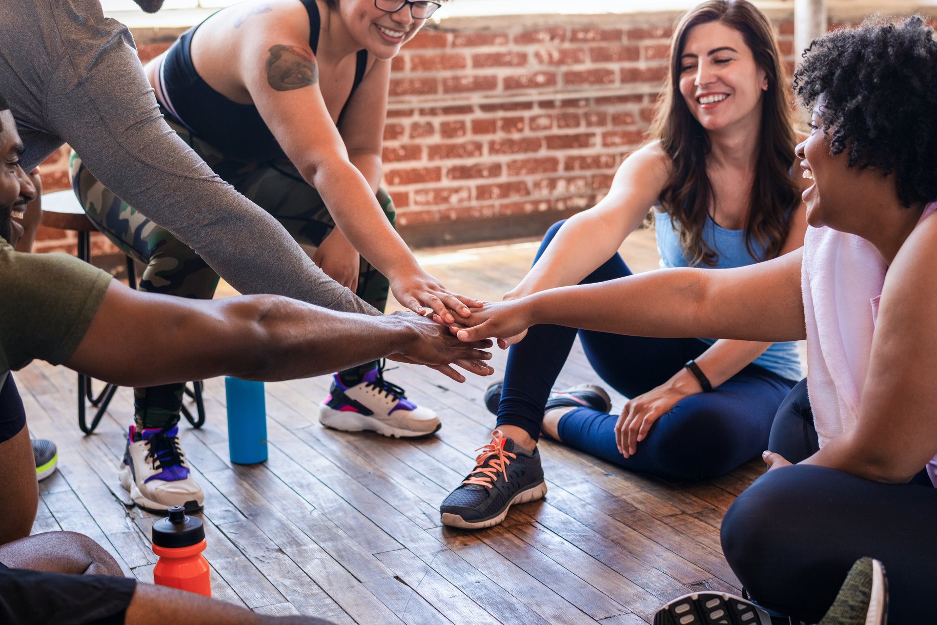 Diverse group of people sitting in circle, hands together in the center, showing teamwork and unity in a casual setting with wooden floors and brick walls. Diverse group of people exercising together