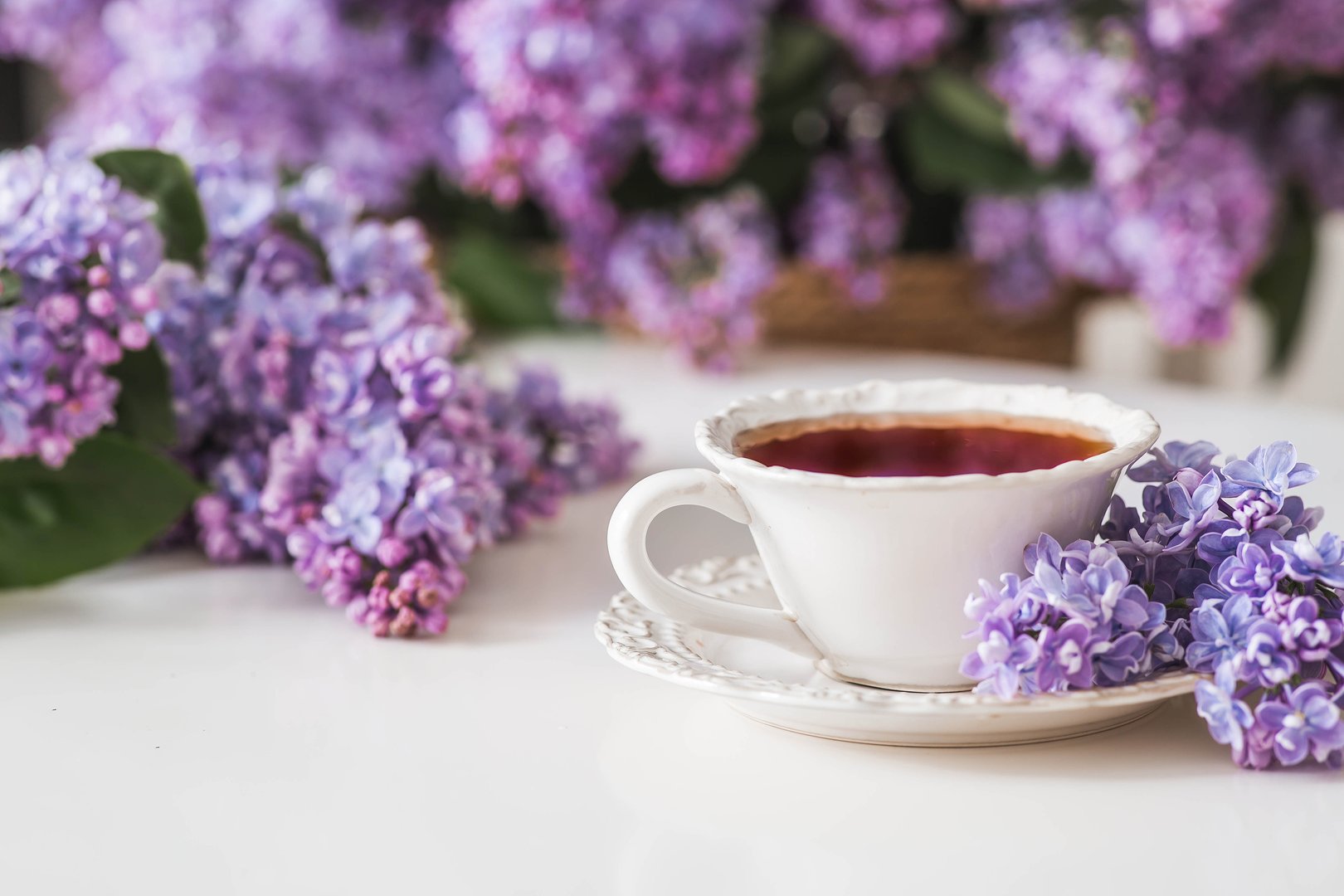 A cup of tea or coffee on the table. In the background is a large purple bouquet of lilacs. The concept of home comfort and drinks.