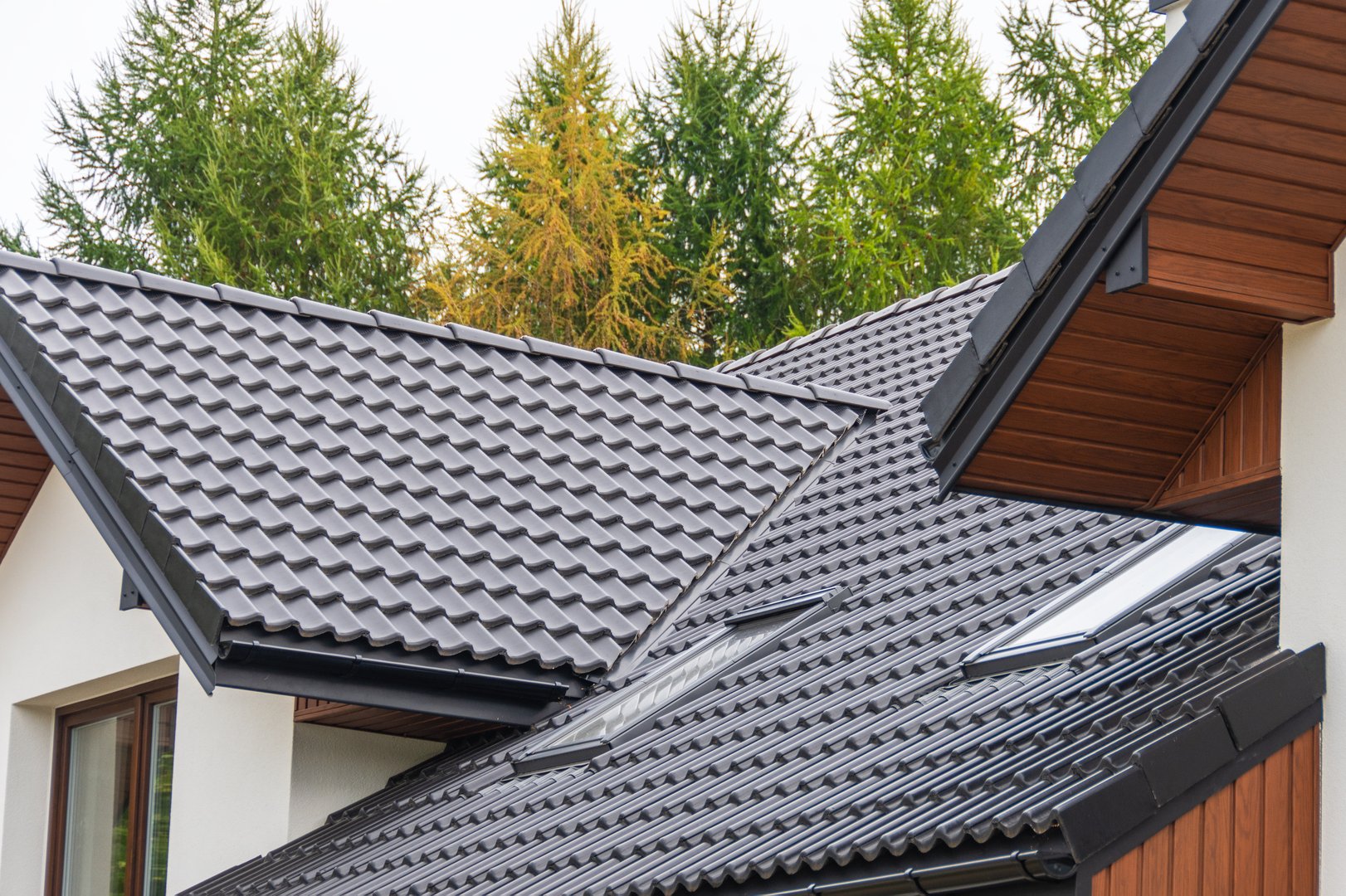 Stylish multi-pitched roofs with dark tiles and wooden accents contrast beautifully with vibrant greenery, showcasing contemporary home design and construction in a suburban setting.