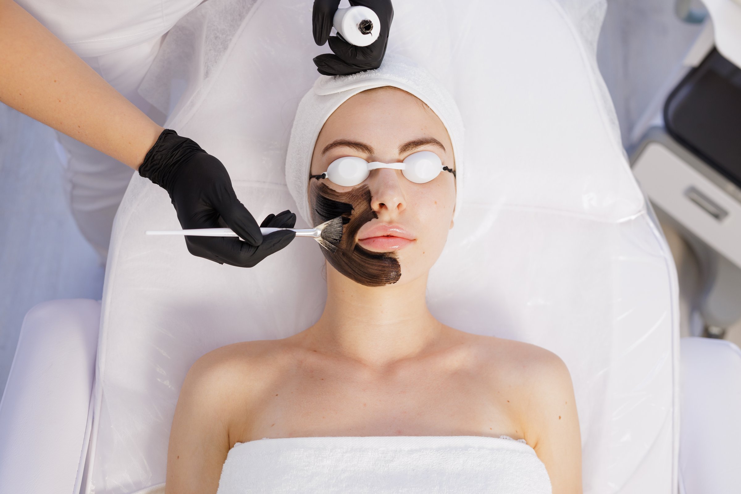 Gloved beautician applies black peel to a woman with eye shields in a bright clinic for exfoliation, cleansing, and glow.