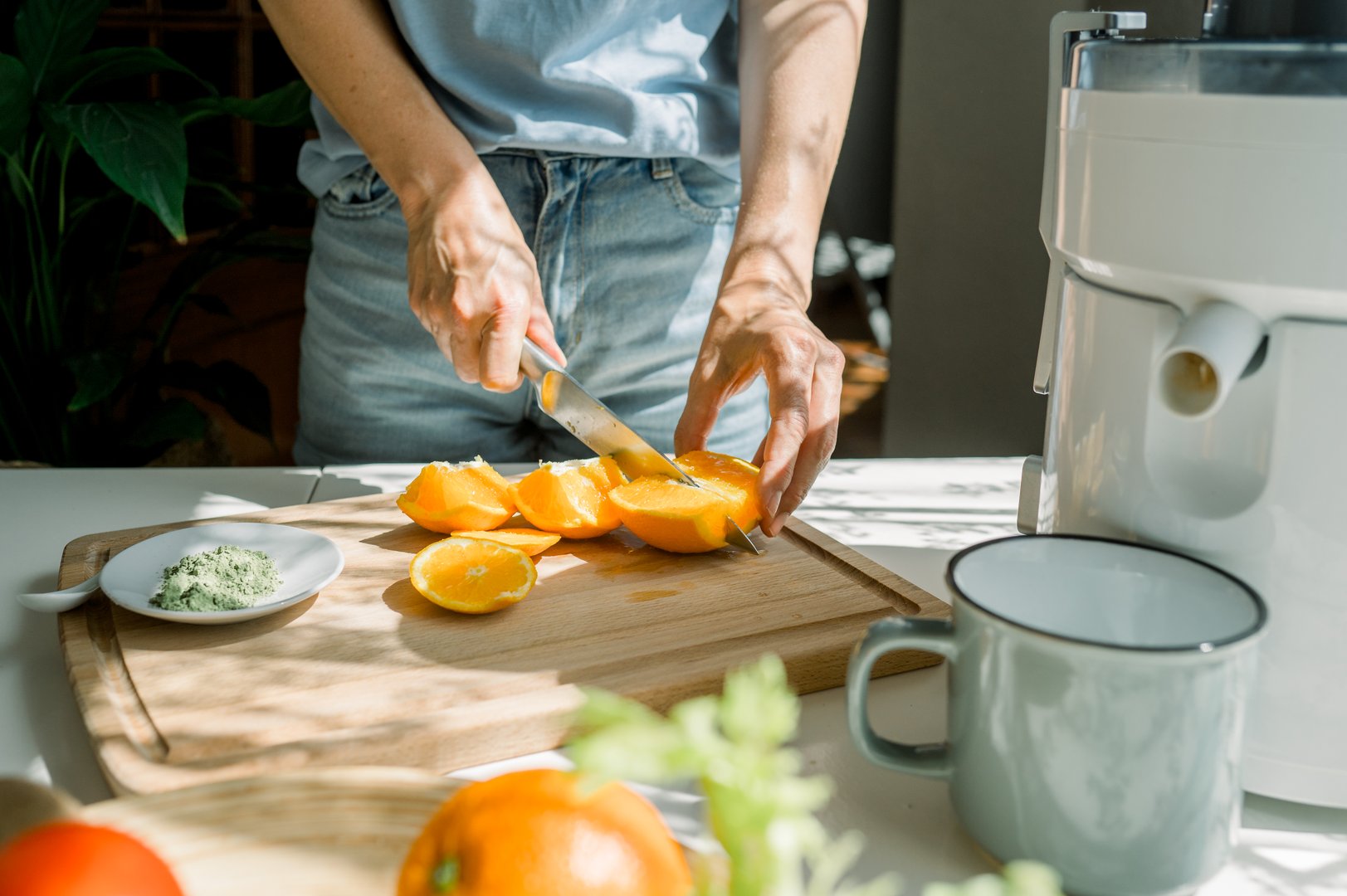 Crop anonymous housewife cutting fresh orange on wooden chopping board while making citrus juice with juicer in kitchen during sunny day