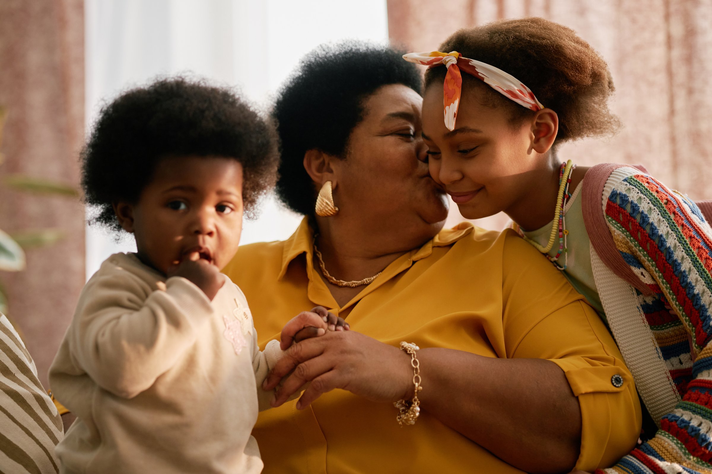 Grandmother gently kissing grandchild on cheek while embracing other grandchild, showing tender affection within cozy home setting