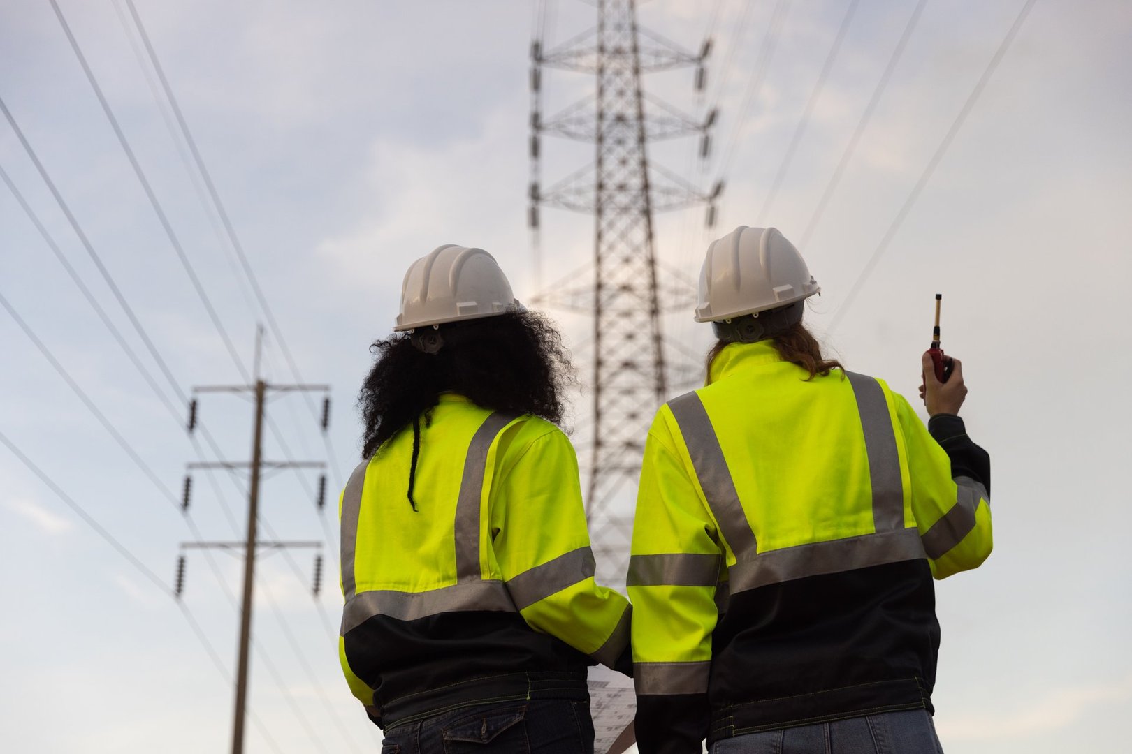 Back view of two female industry engineers working and discussing in front of a voltage-generating station and  high voltage pole